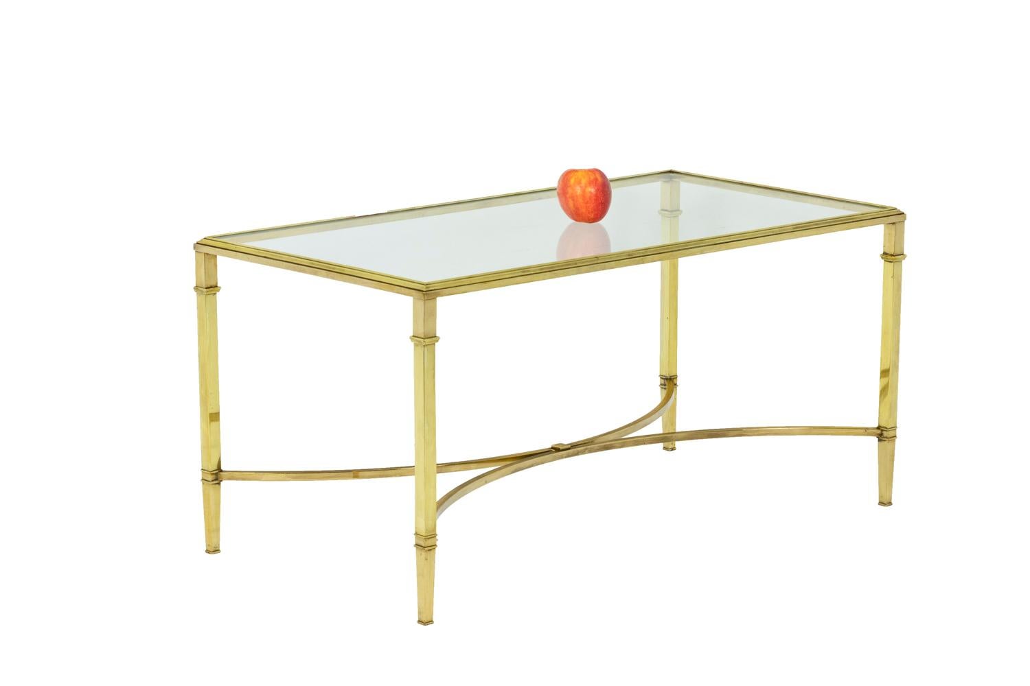 Lounge coffee table in solid and gilt bronze. The rectangular top is composed of a molded frame that fits a transparent glass. The Four square section and slightly tapered feet in the lower part are adorned with rings and connected by an X-shaped