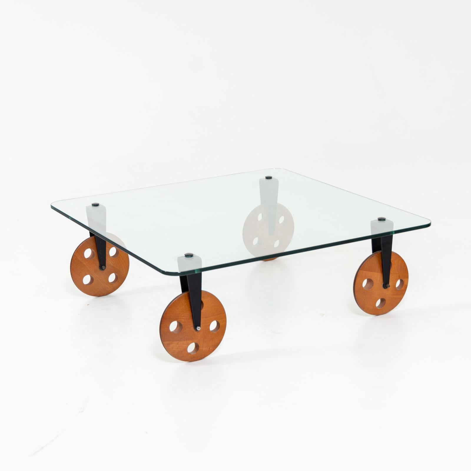 Quadratischer Couchtisch mit Glasplatte im Stil von Gae Aulenti. Der Tisch steht auf Holzrädern, die über schwarze Metallbeschläge mit der Platte verbunden sind.