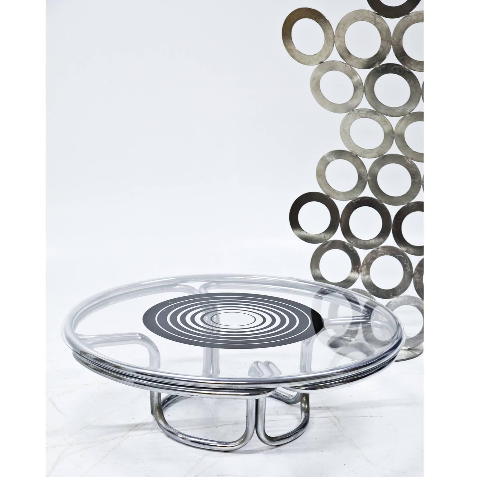 Coffee table on a bent and chromed metal frame. The round tabletop has some black circles printed on it and is made out of plastic.