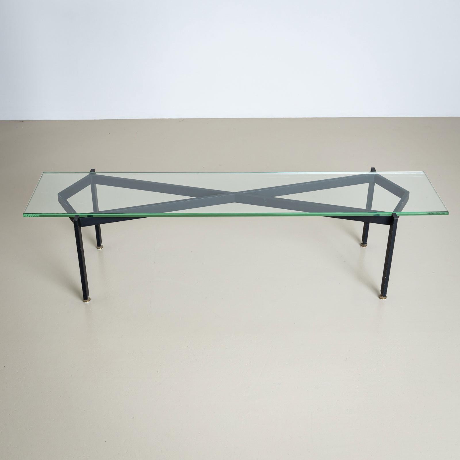 Table basse rectangulaire avec un plateau en verre transparent reposant sur un cadre en fer noir en forme de X. La table repose sur des pieds droits avec des pieds décoratifs en laiton. La finition laquée noire présente quelques zones d'usure, ce