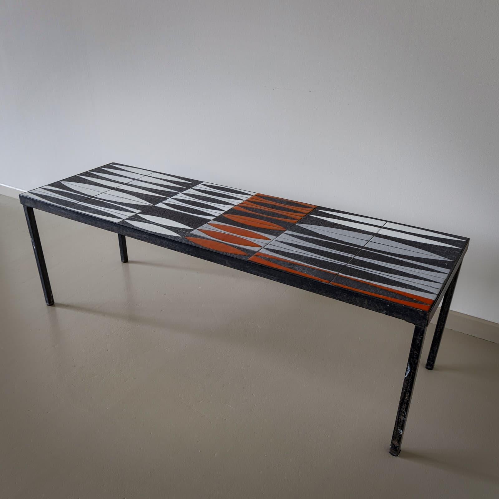 

Mesa de centro con estructura de hierro lacado en negro y tablero rectangular de azulejos de cerámica. Estos azulejos presentan un dibujo geométrico en negro, blanco y rojo. El tablero está firmado 