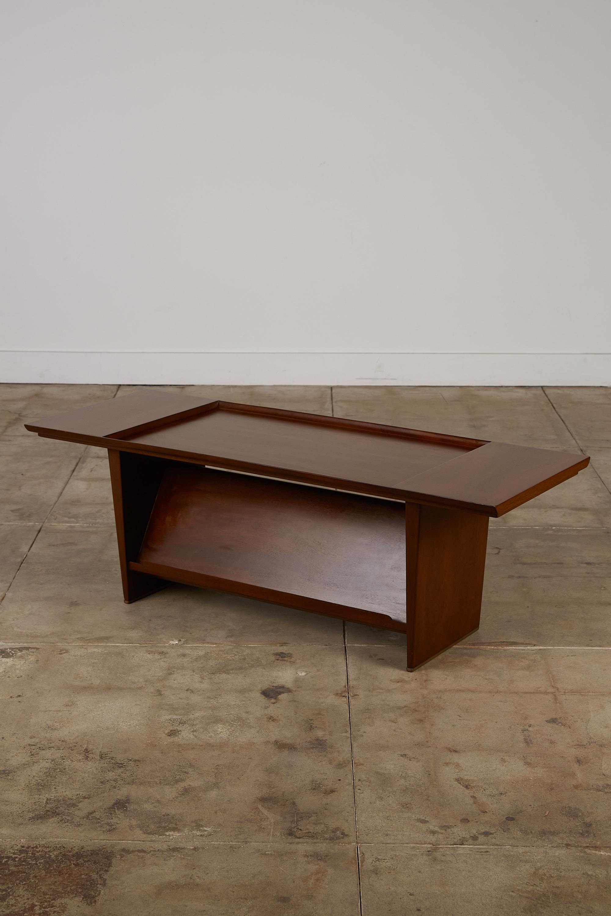 A mahogany coffee table for home or office, with a built-in display shelf, designed by Edward Wormley for Dunbar. The understated design has a rectangular top with a recessed well in the centre. Two mahogany panel trestles support the tabletop and