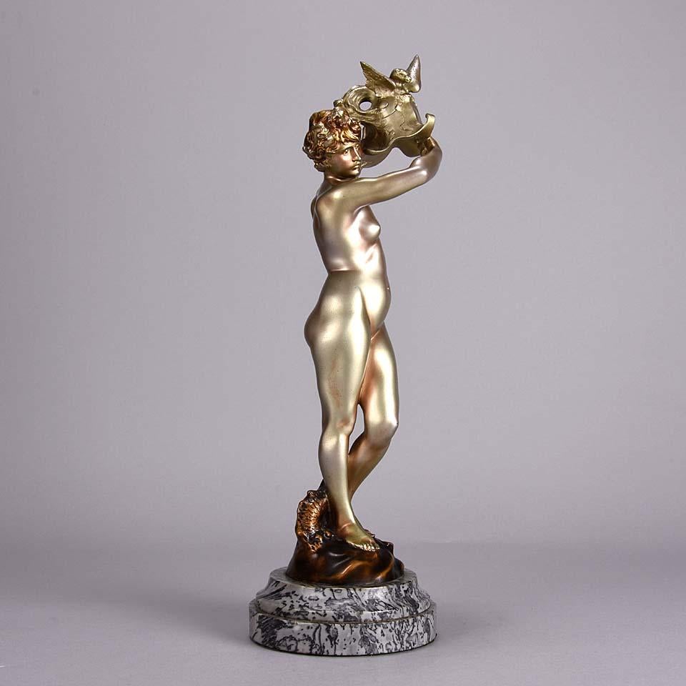 A fabulous early 20th century Art Deco cold painted silver bronze figure of a beautiful woman standing naked on a naturalistic mound as she holds aloft a winged dragon helmet with a dragon wrapped around her legs. The bronze surface with excellent