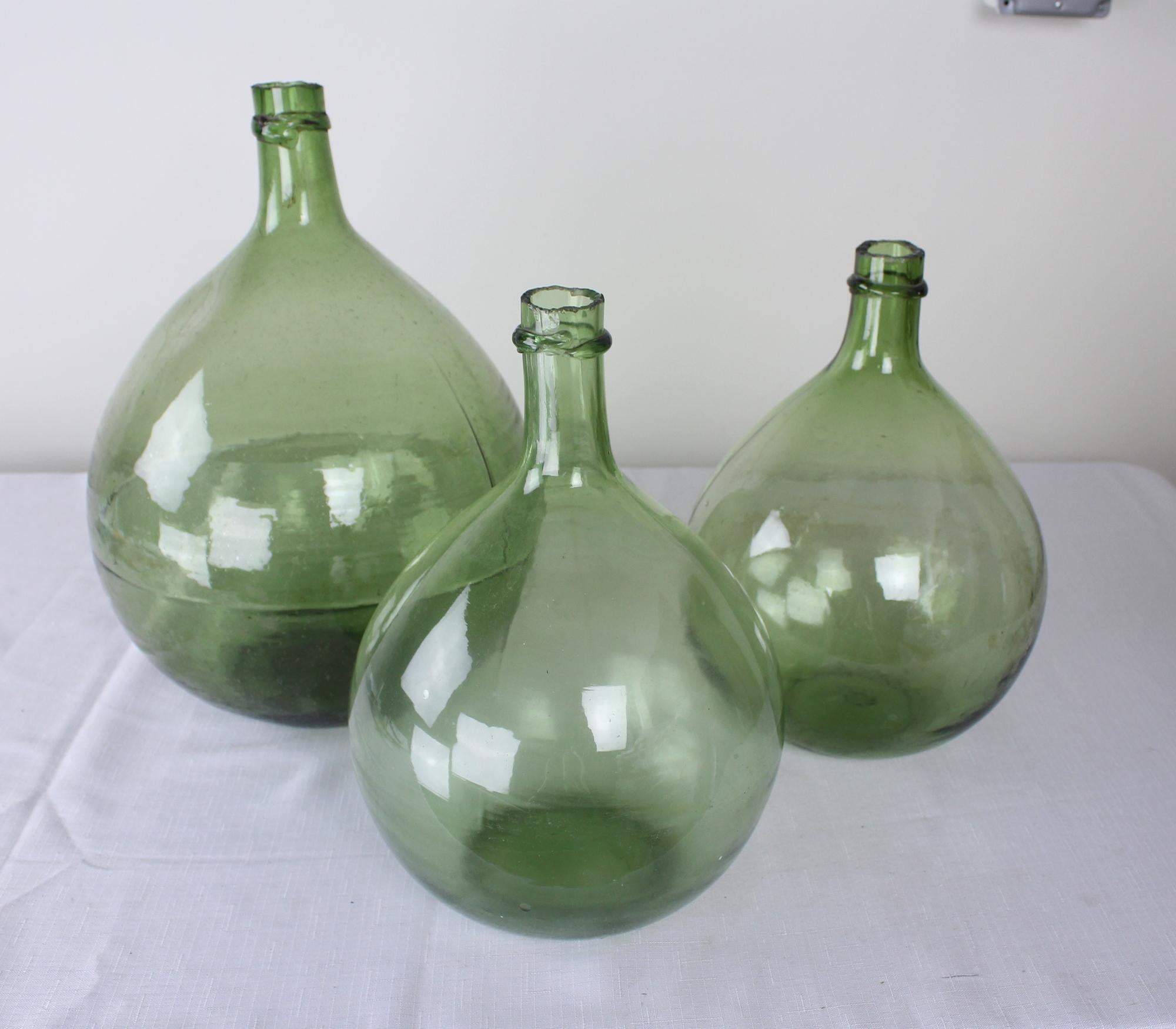 A collection of three olive colored glass demijohn bottles, used from the late 17th century through the 19th century to carry wine and spirits. Hand blown, each has their own unique characteristics, from the tilt of the gooseneck to the trapped air