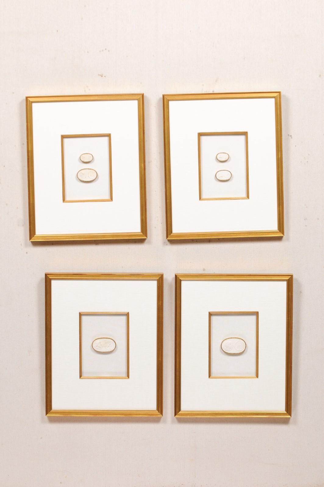 A collection of four framed assortment of Greek intaglios in white, set in custom gold frames. This set of wall art features hand-cast intaglios (imprints from the original ancient coinage castings, likely European in origin), ovoid in shape with