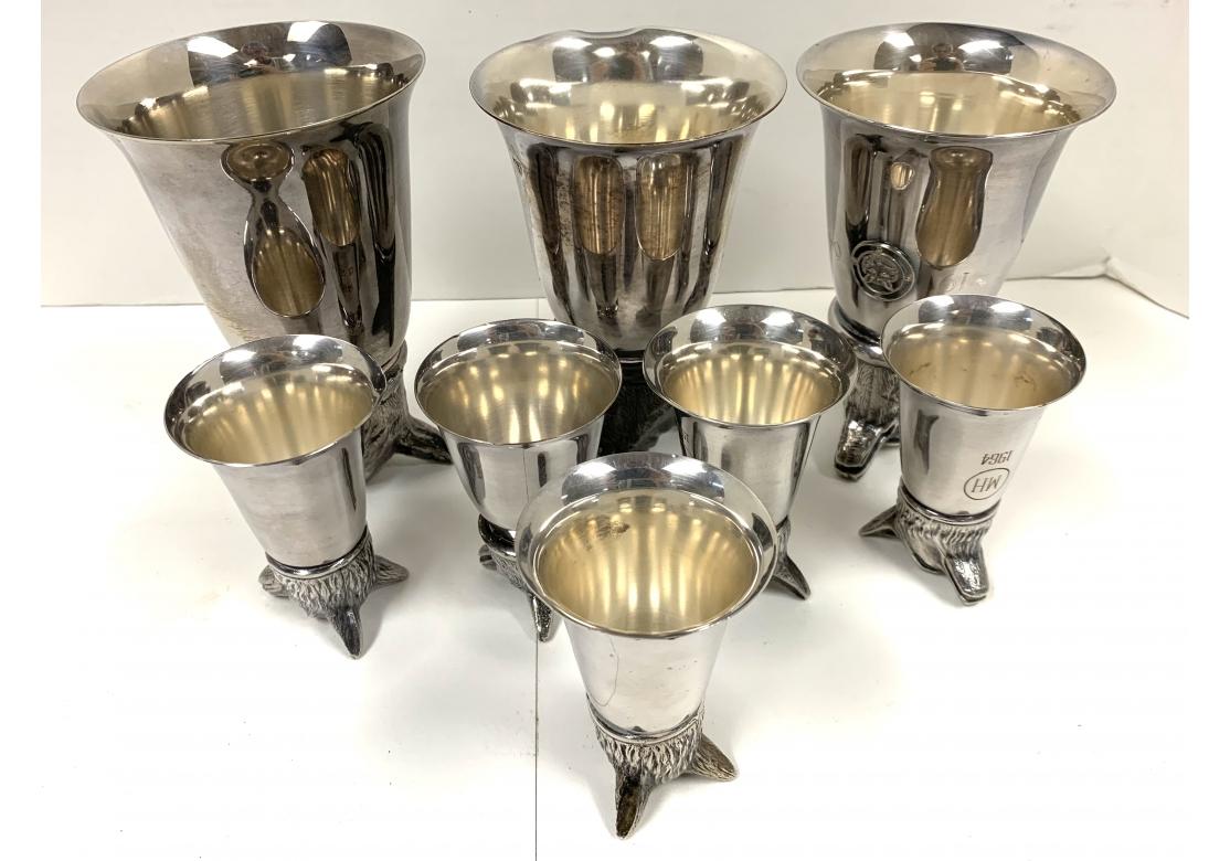 Collection of 8 vintage metal stirrup fox head trophy cups in two sizes with monograms and dates. The three larger pieces are not identical but compatible. One with fox medallion on front dated 1964 and hallmarked, similar one also dated 1964 and