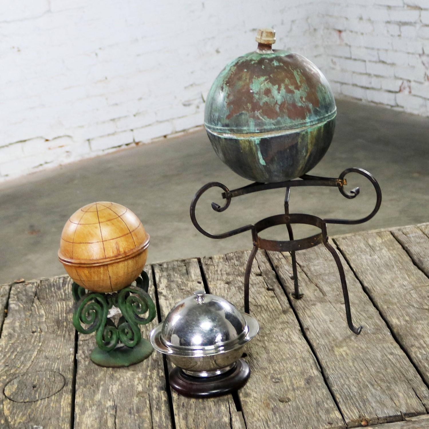 Fun set of three orb shaped object on stands to be used as centerpiece or object d’art. Large copper orb on wrought iron stand, carved wood globe box on cast iron stand, and chrome round lidded compote on a flat wood stand. They are all in wonderful