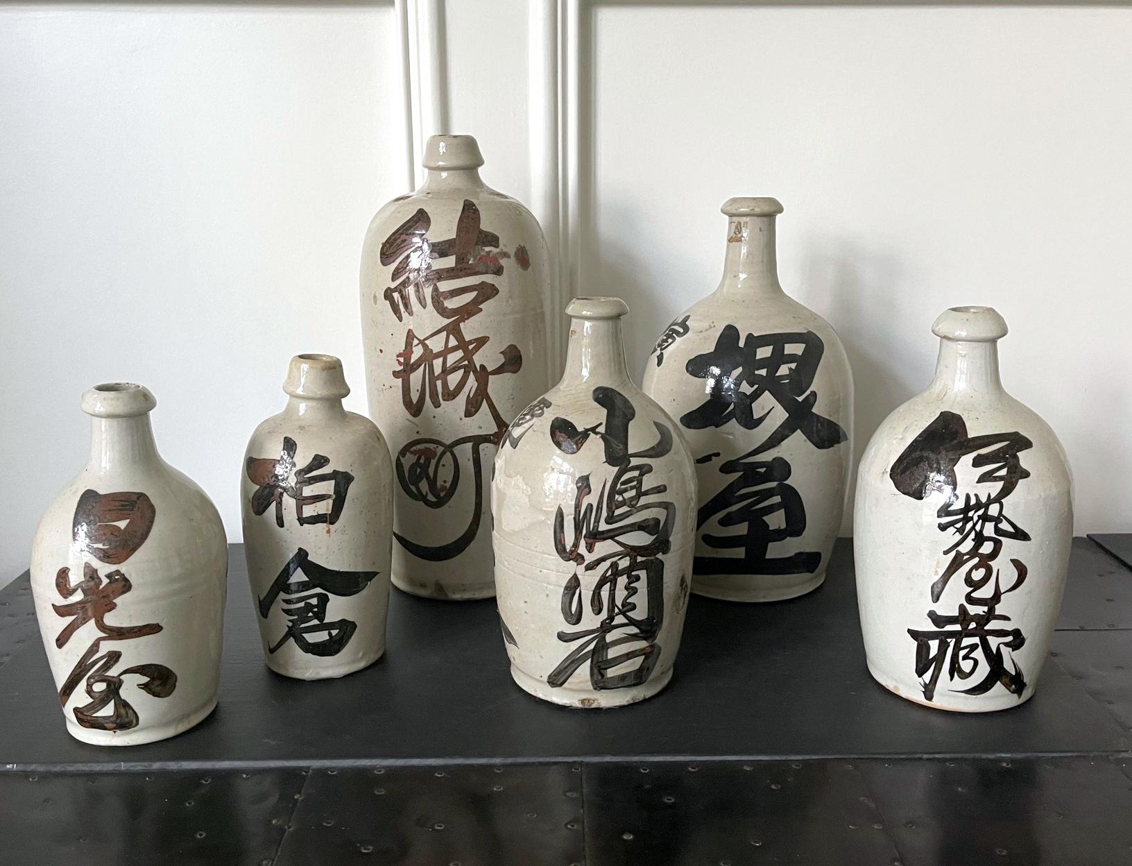 A collection of six Japanese stoneware sake bottles circa first quarter of 20th century. Heavily potted, these bottled were covered in white glaze and brushed on with black Kanji characters to show the brand names of sake or the wine houses. They