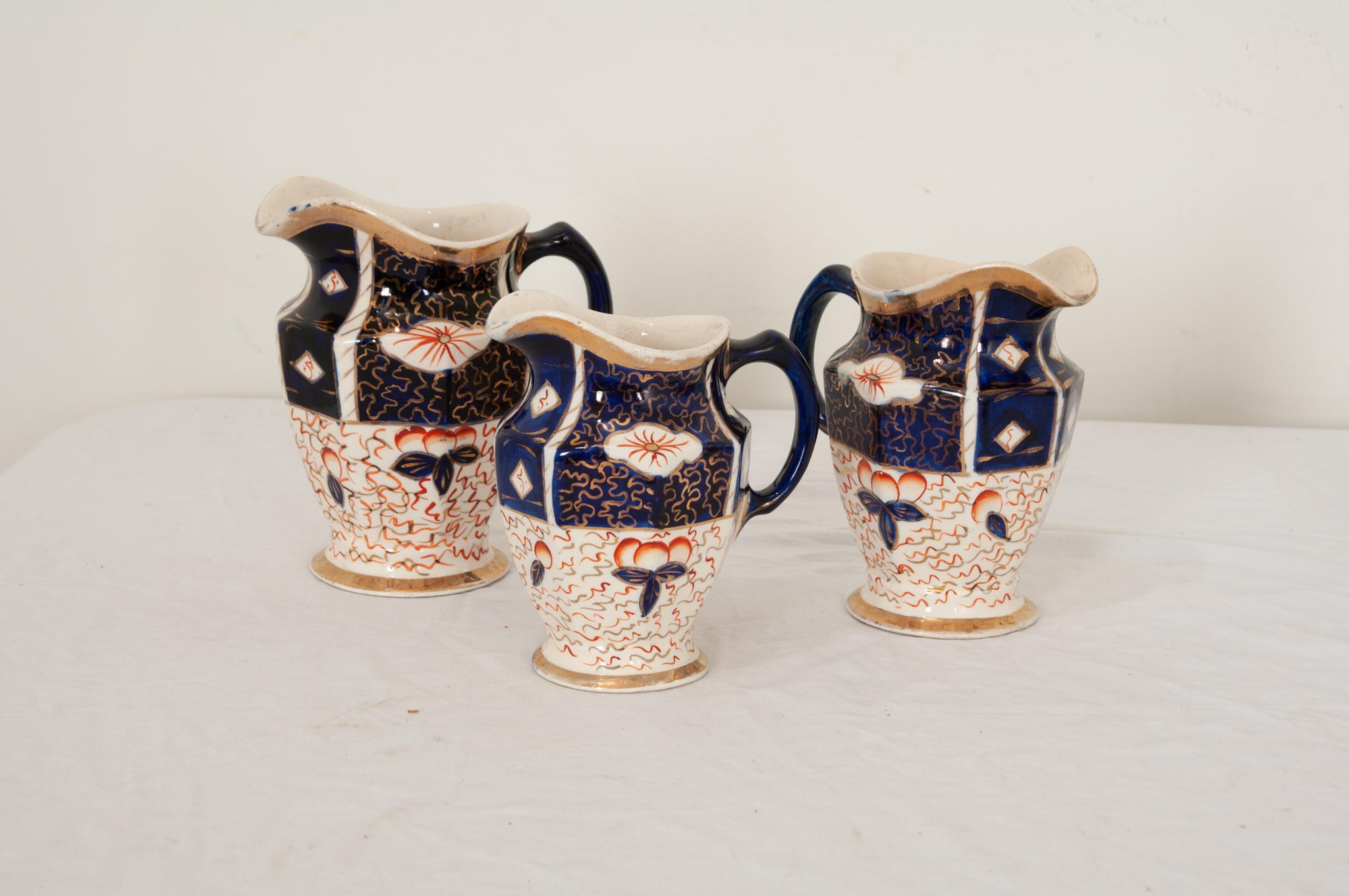 A collection of English 19th century porcelain pitchers with a hand painted finish. Sold as a set, these decorative pitchers feature rich cobalt blue and deep red Asian aesthetic designs on creamware that offsets the hand painted gold highlights.