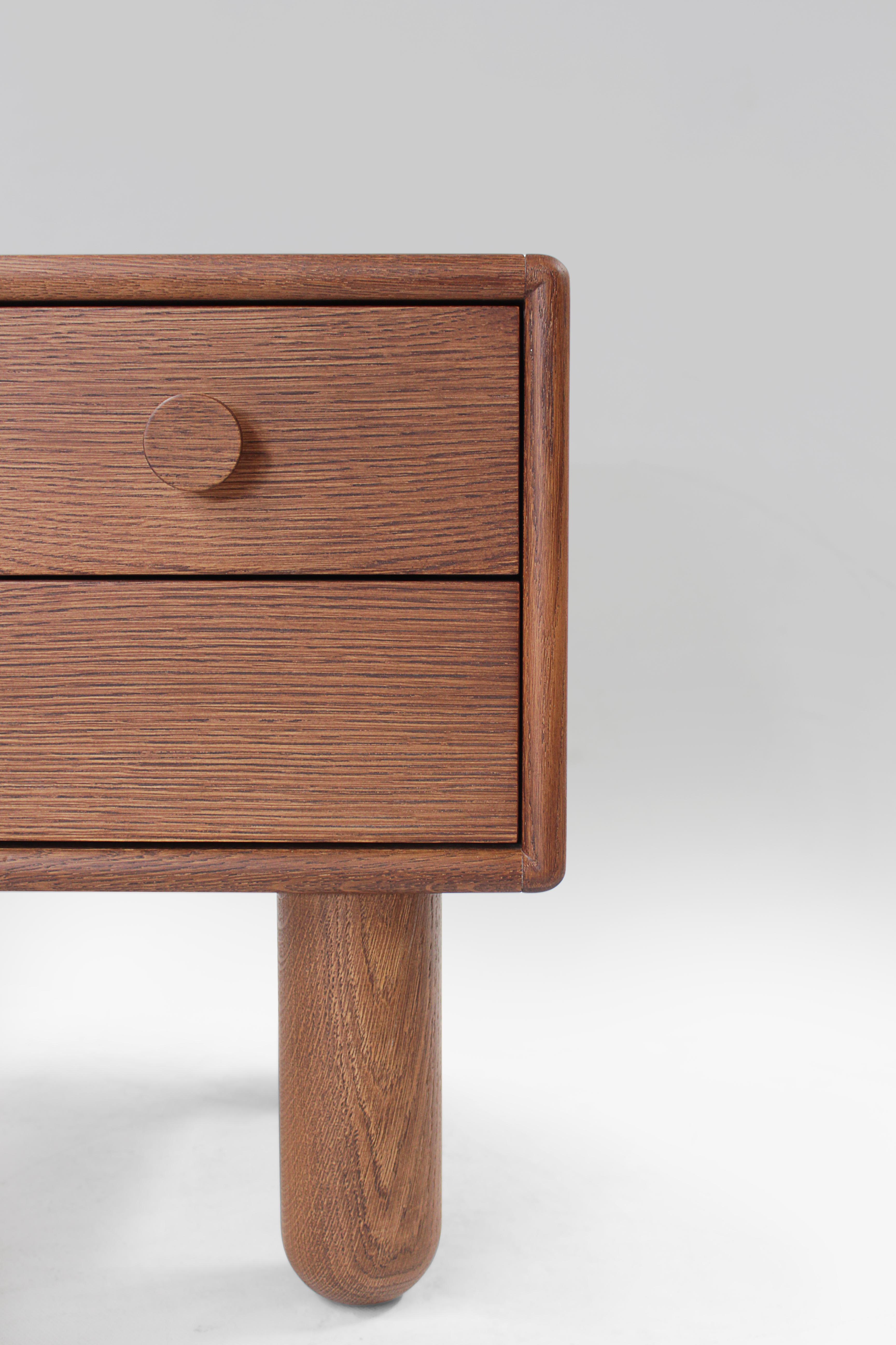 Table de chevet en bois de belle facture, au design minimaliste et aux arêtes douces et arrondies. Il comporte trois tiroirs avec des boutons circulaires en bois, offrant un rangement pratique tout en conservant une esthétique élégante. Soutenue par