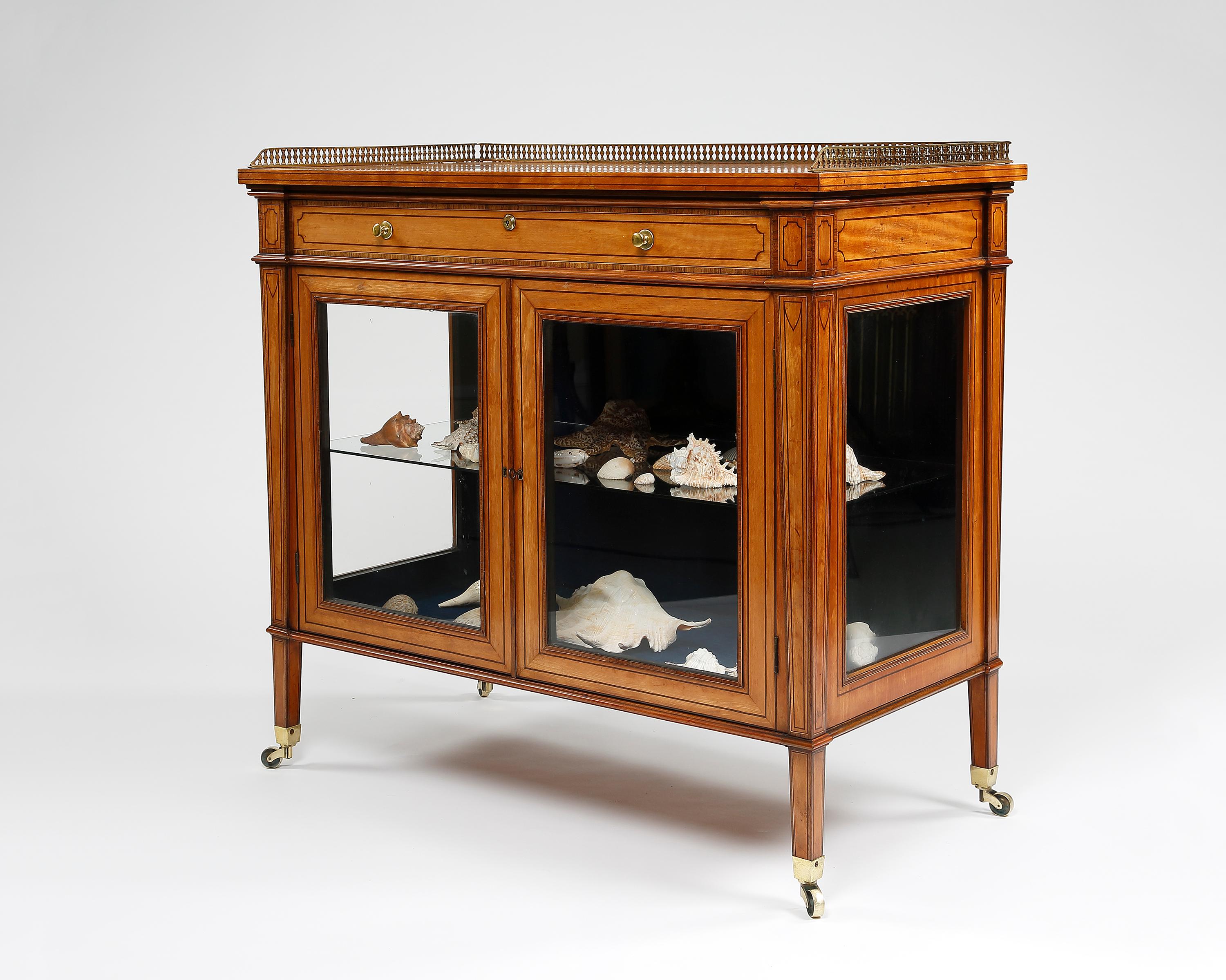 A fine quality two-door satinwood glazed door cabinet with glazed sides, and original brass gallery. Below is a single cedarwood lined drawer with original brahma lock and brass knob handles. The whole is raised on four long square tapering legs