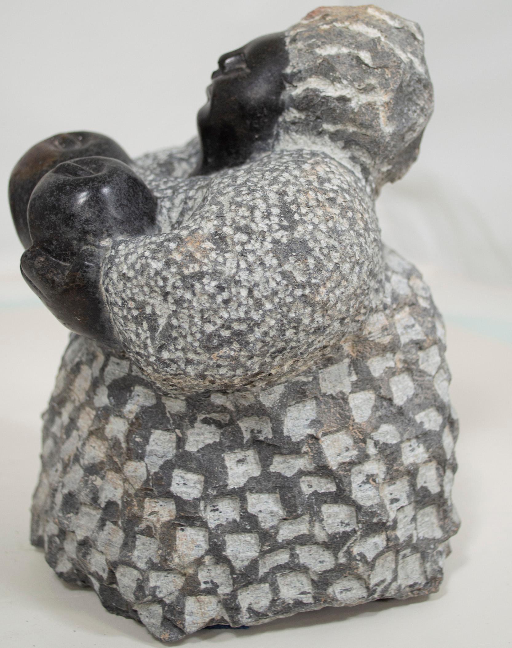 'Holding Apples' is an original black serpentine sculpture by the celebrated second generation Shona artist Colleen Madamombe. The sculpture presents a character common to Madamombe's work: a woman with a round face and wearing a billowing dress. In