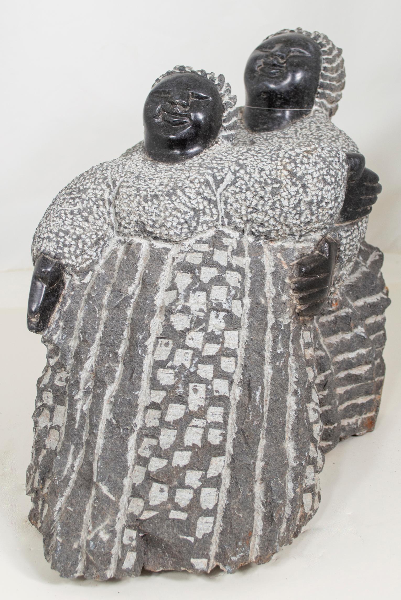 Playing Sisters" est une sculpture originale en serpentine noire réalisée par Colleen Madamombe, célèbre artiste shona de la deuxième génération. La sculpture présente un personnage commun à l'œuvre de Madamombe, à savoir une femme au visage rond,
