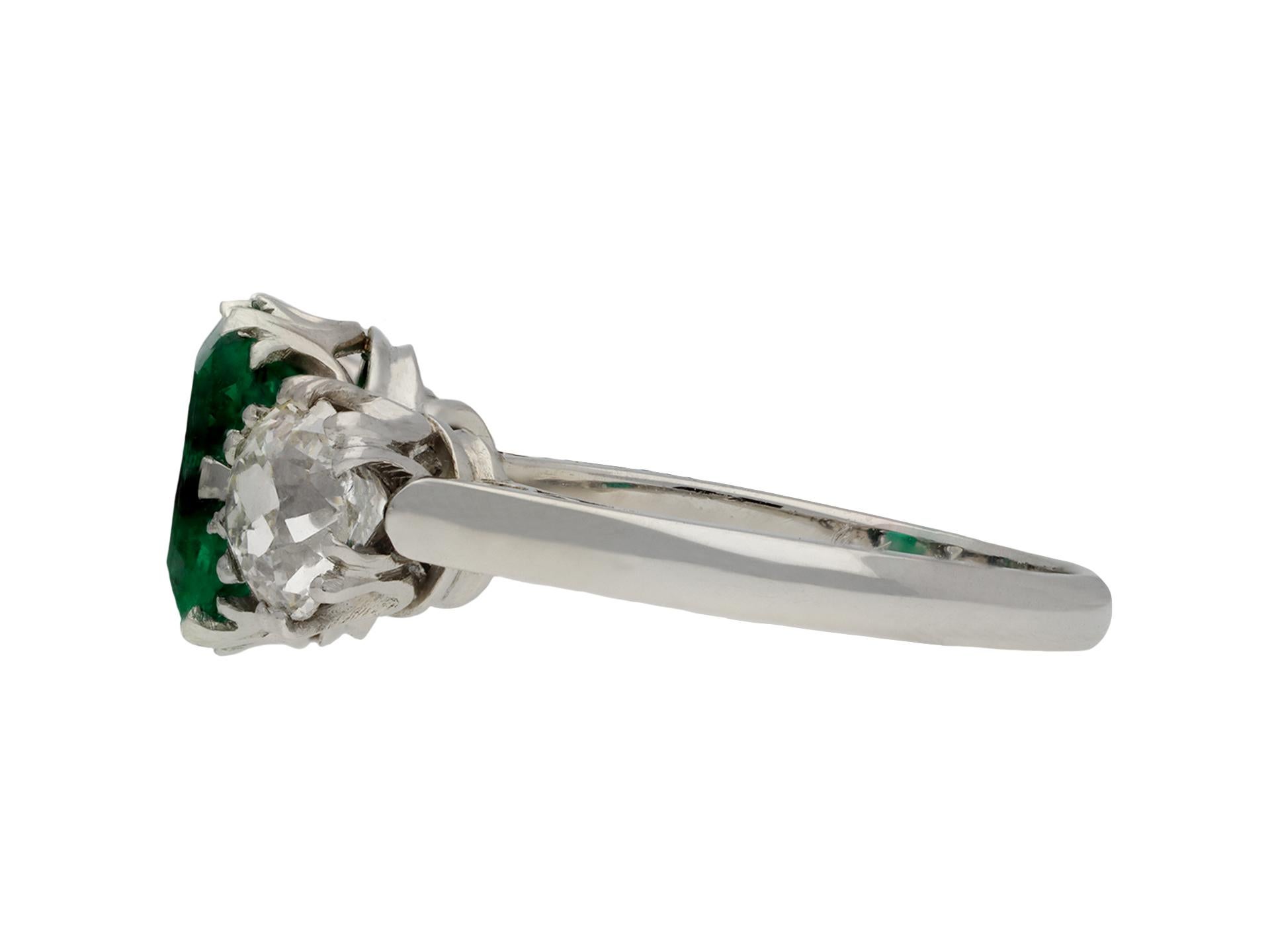 Colombian emerald and diamond three stone ring. Set to centre with a cushion shape old cut natural Colombian emerald with no colour enhancement and minor indications of clarity modification, in an open back claw setting with an approximate weight of