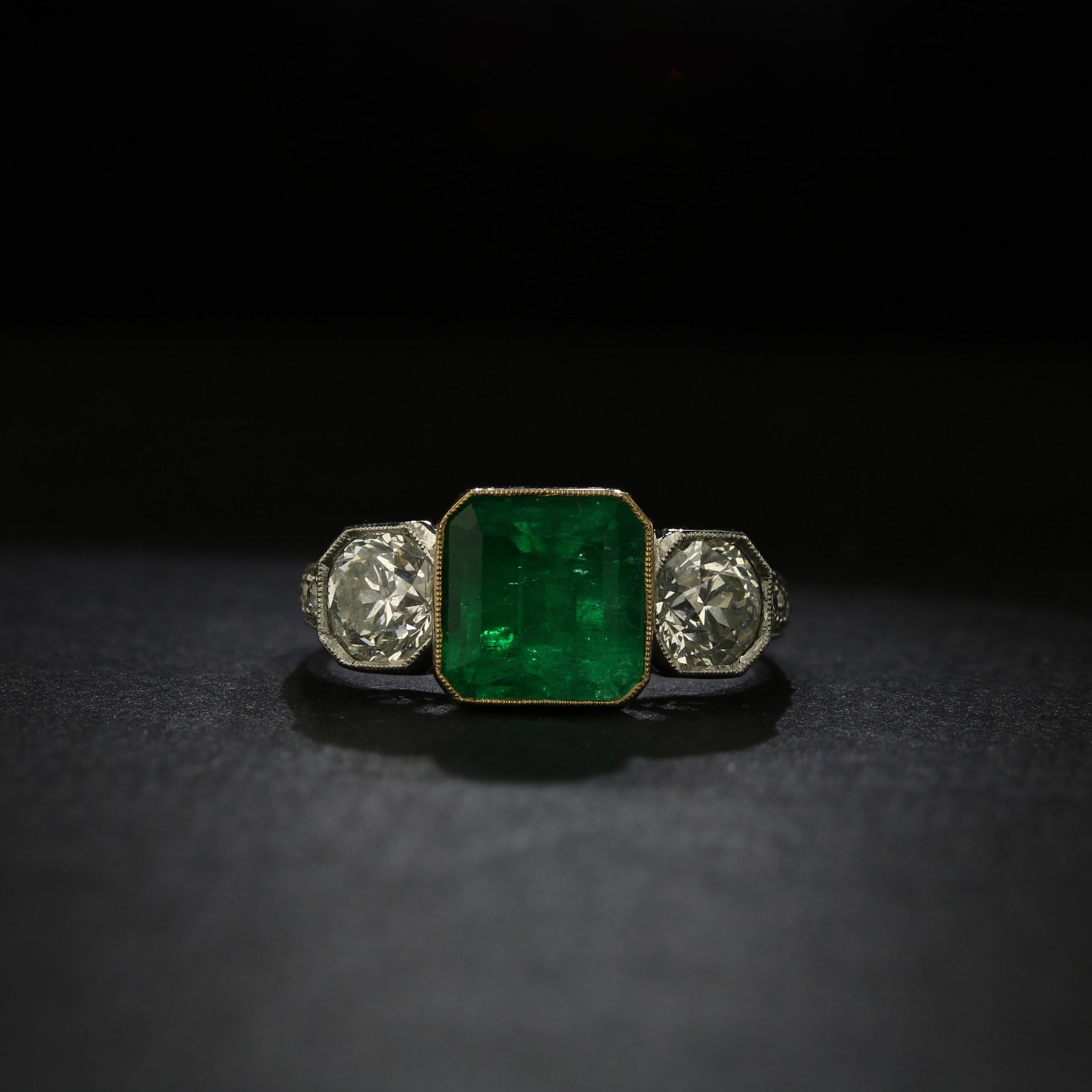 One antique Colombian emerald and diamond ring. Crafted in platinum, the ring features a square-cut green emerald of approximately 2.50 carats, bezel set in the center with yellow gold accent. On either side of the emerald is an old European