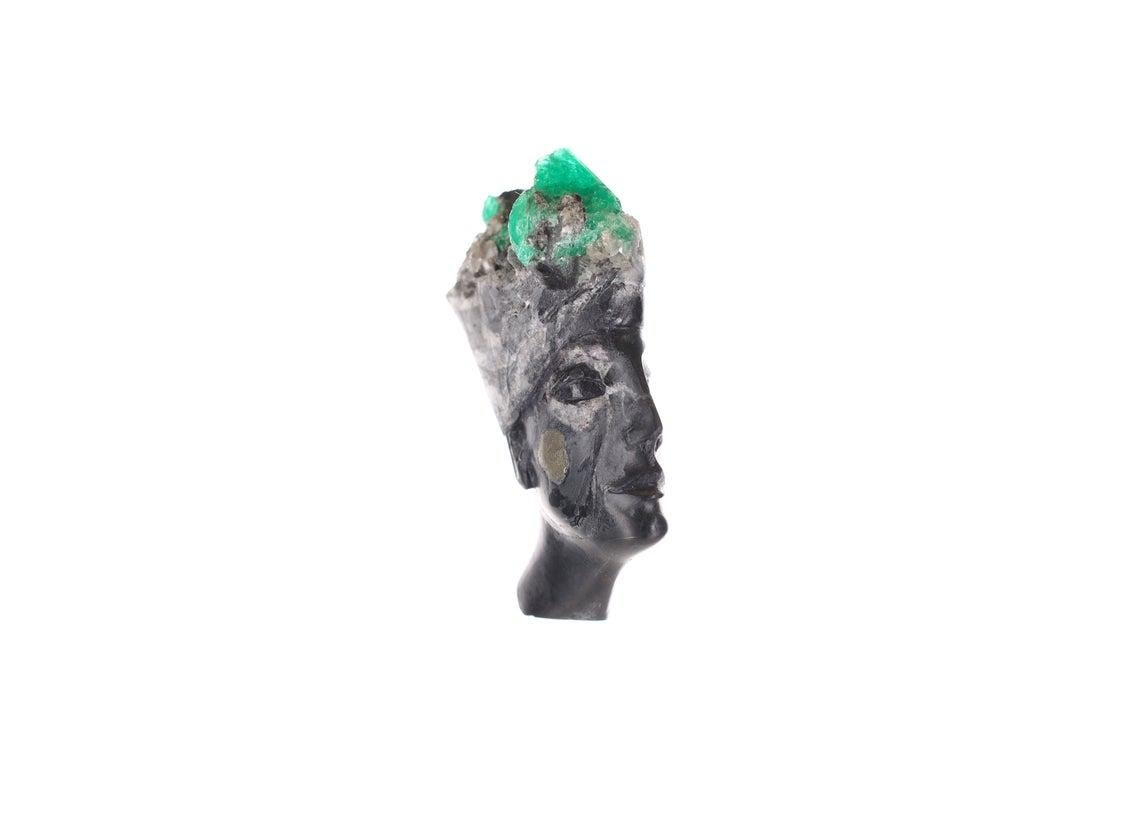 This is a beautiful and one-of-a-kind rough Colombian emerald sculpture. Featuring a magnificent Egyptian goddess, made of black and gray shale with bits of calcite and a patch of pyrite on the right side of her face. Her crown is made up of natural