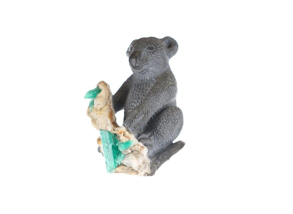 This beautiful and one-of-a-kind rough Colombian emerald sculpture features a hand-carved Koala made of gray shale. It is holding a piece of rock containing rough Colombian emeralds, white calcite, and pyrite. The detail of this piece is quite