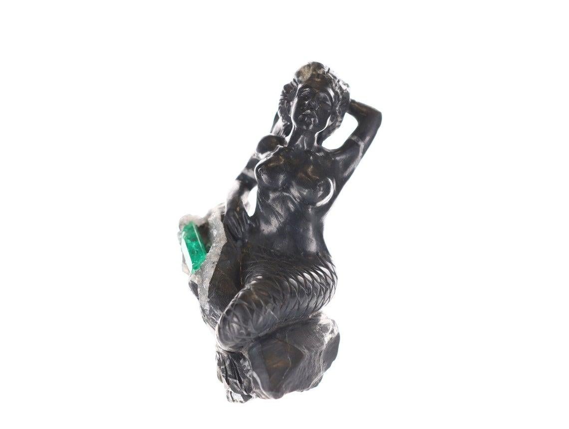 This is a beautiful and one-of-a-kind rough Colombian emerald sculpture. It displays a gorgeous hand-carved mermaid of black and gray shale, with intricate detail from the scales of her tail to the hands within her hair. She is sitting on the edge