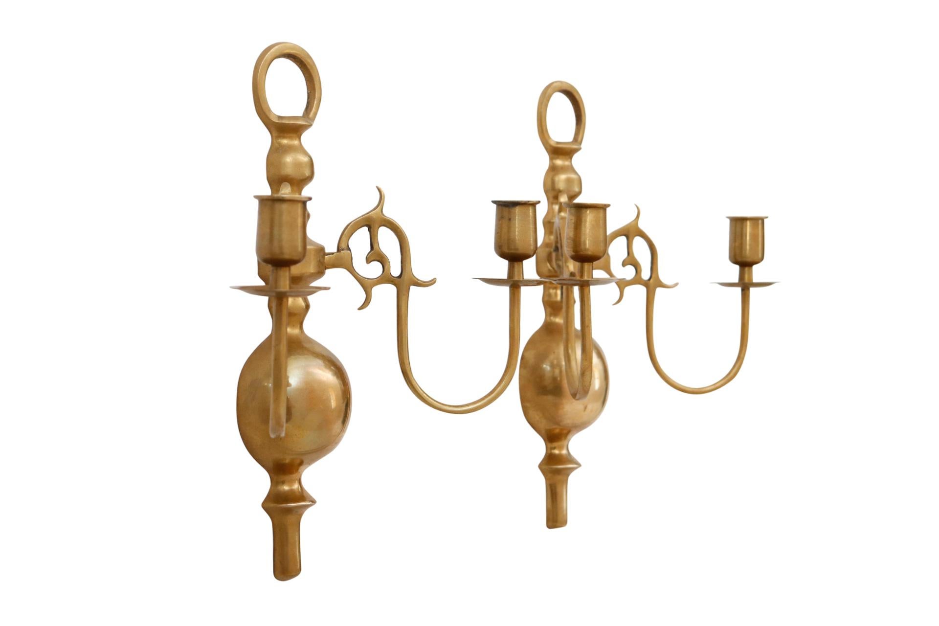 A pair of colonial style brass candle sconces each with two arms. Decorative baluster shaped backplates and hooped tops support two slender arms with scrolled backs. Finished with sleek capitals and bobeches. Dimensions per sconce.


