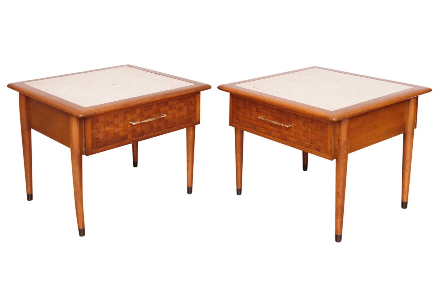 A pair of Colonial style travertine top side tables. Each has a single drawer that opens with a sleek brass handle. Drawer fronts are decorated with woven bamboo. Table legs taper and are finished with brass sabots. Dimensions per table.