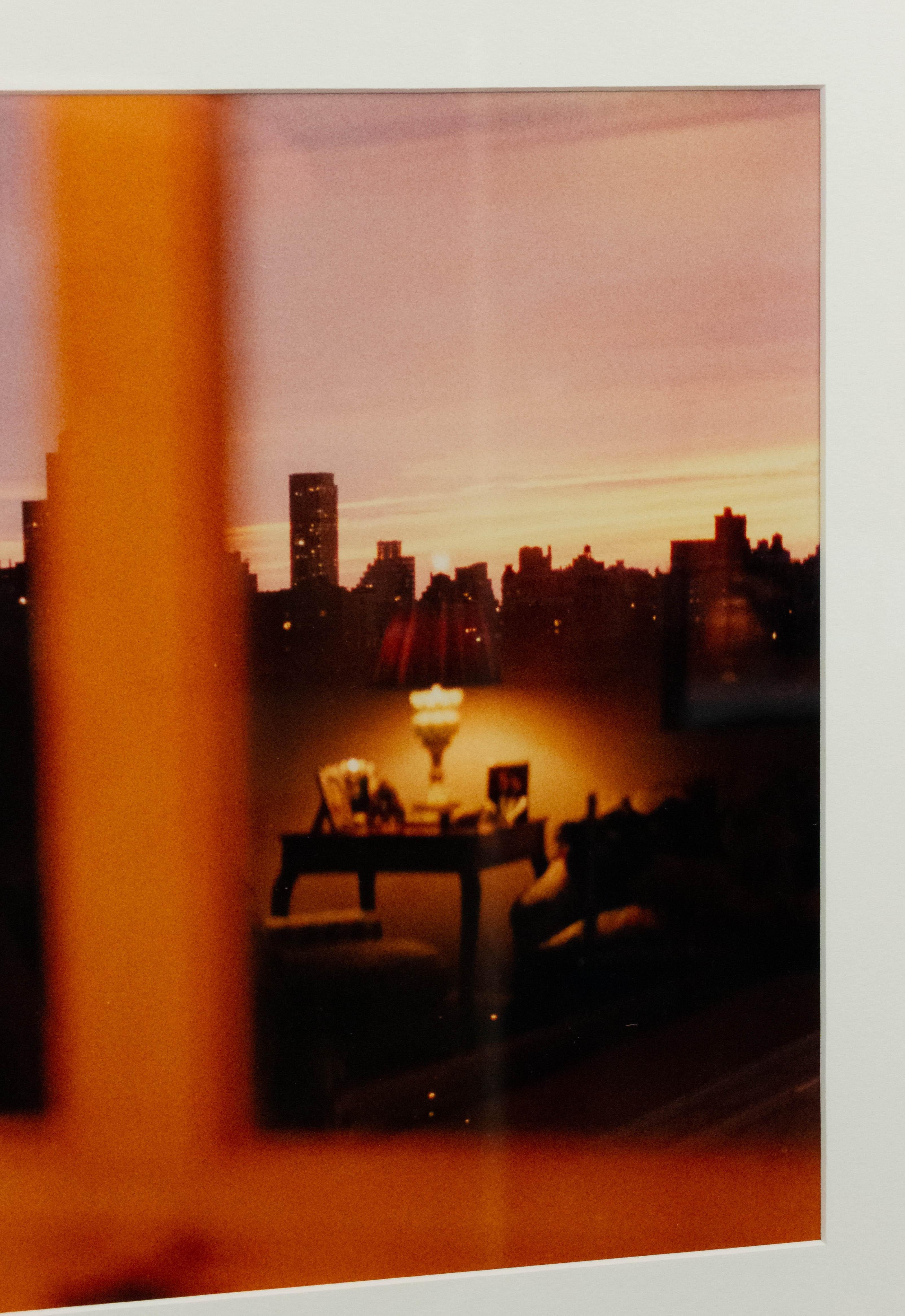 Color photograph of a reflection in a window of a living room and a cityscape with a black frame and a white matte. Photograph by Shelley Harrison.
 