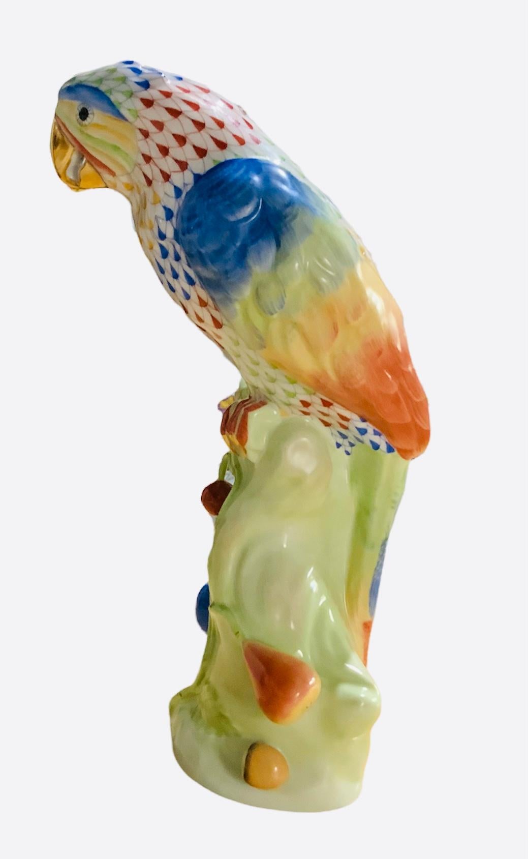 This is a hand painted Herend porcelain parrot. Its background is white and adorned with tropical colors of orange, blue, green, & yellow fish scale net pattern. Also it’s feathers follow the same colors. It is standing over a wood branch that have