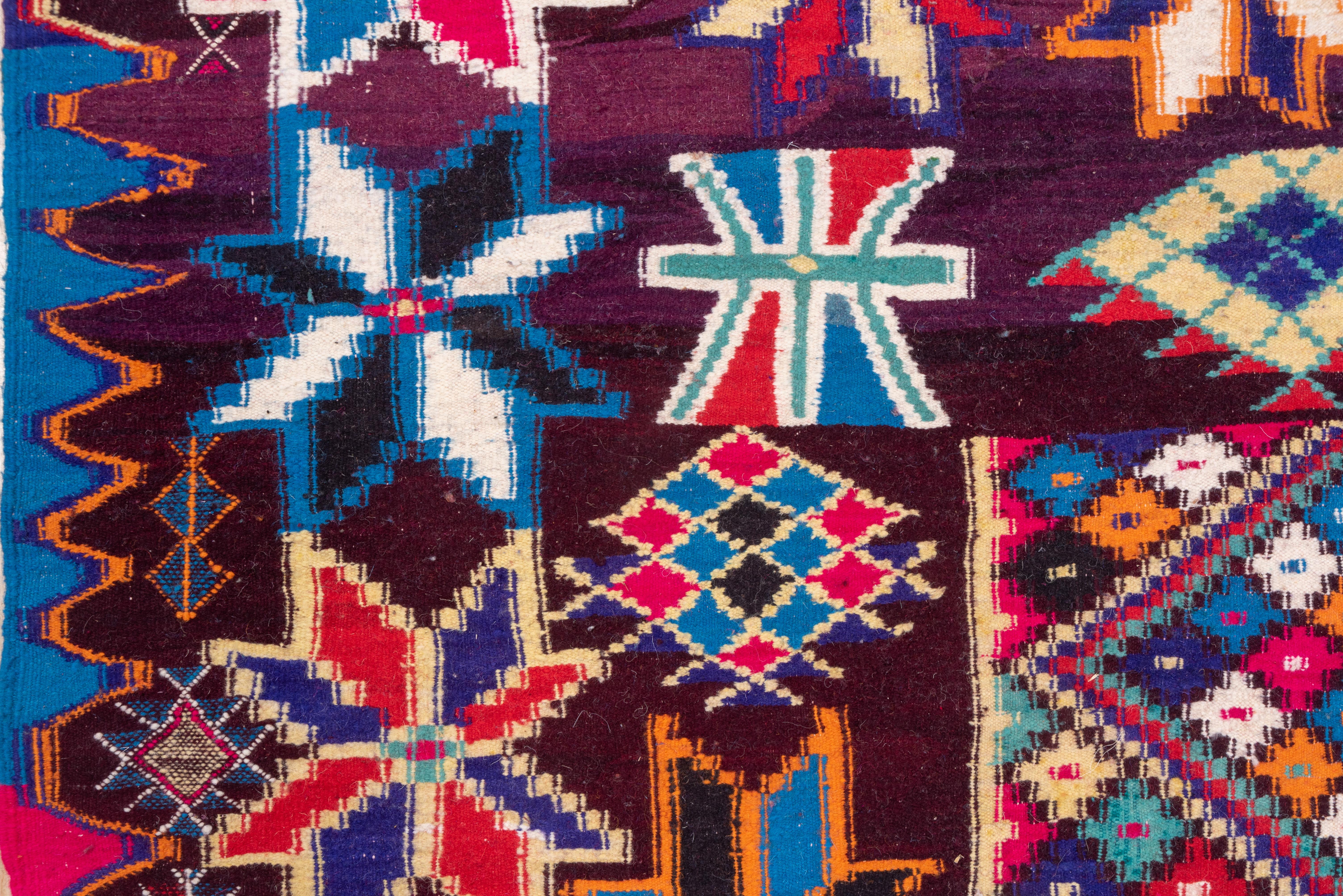 The eggplant ground of this tribal flat-weave carpet displays eleven rows of colorful stars set closely around a small central rectangle with a polychrome lozenge mosaic. At one end is a border of double triangles while at the other is a partial
