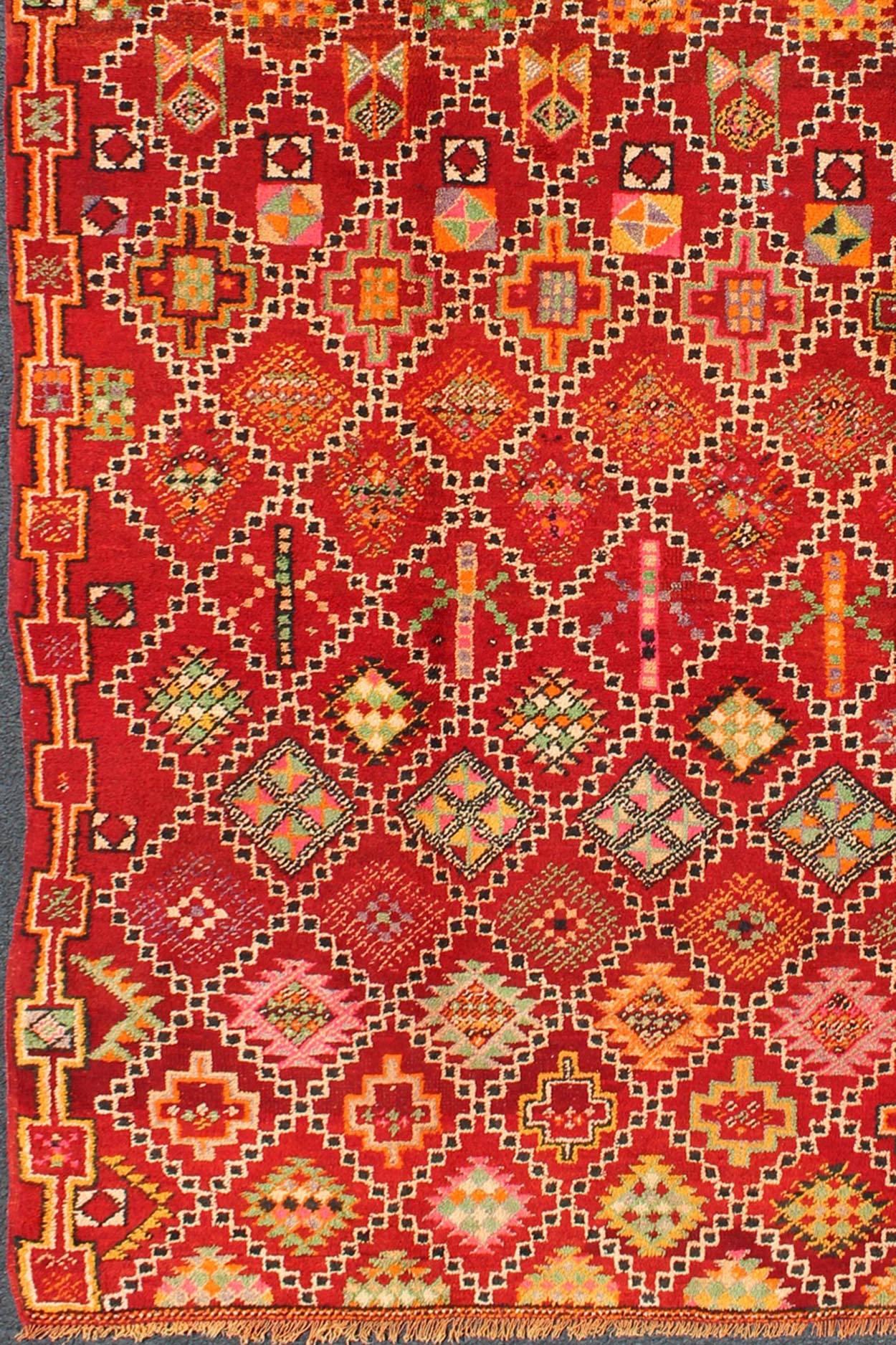 This gorgeous Vintage Moroccan carpet from the mid-20th Century displays a stunning, all-over geometric pattern and palette of rich colors. The red base of the rug holds a black & white diamond-shaped grid pattern that contains a wide variety of