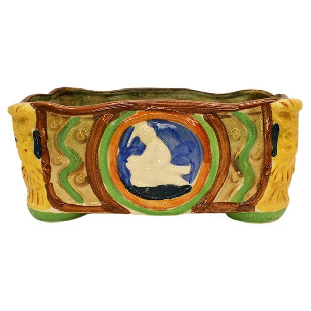 A beautiful colorful rectangular Japanese majolica planter. Each corner features a lion head motif in bright yellow. Each of the sides features a figurative medallion in red, maroon, blue, and cream. The bottom features bright green glazed footing.