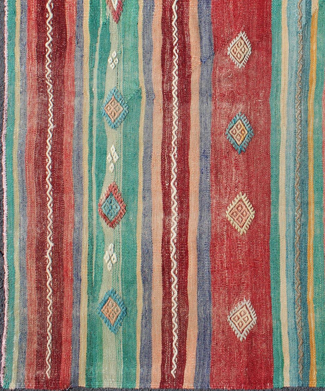 Woven during the mid-20th century in Turkey, this designer Kilim is decorated with a vertical stripe pattern with geometric designs throughout. This flat-woven Kilim is rendered in shades of red, orange, greens, blues and ivory. This rug is