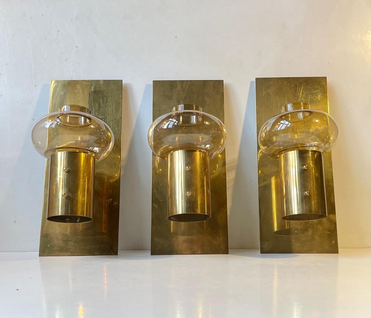 Attractive trio of mid-century wall candle lamps - sconces in solid brass made by Colseth Norway during the 1960s. Featuring tulip shaped smoky amber colored glass shades placed in front of the simple yet stylish back panels. Removable glass. For