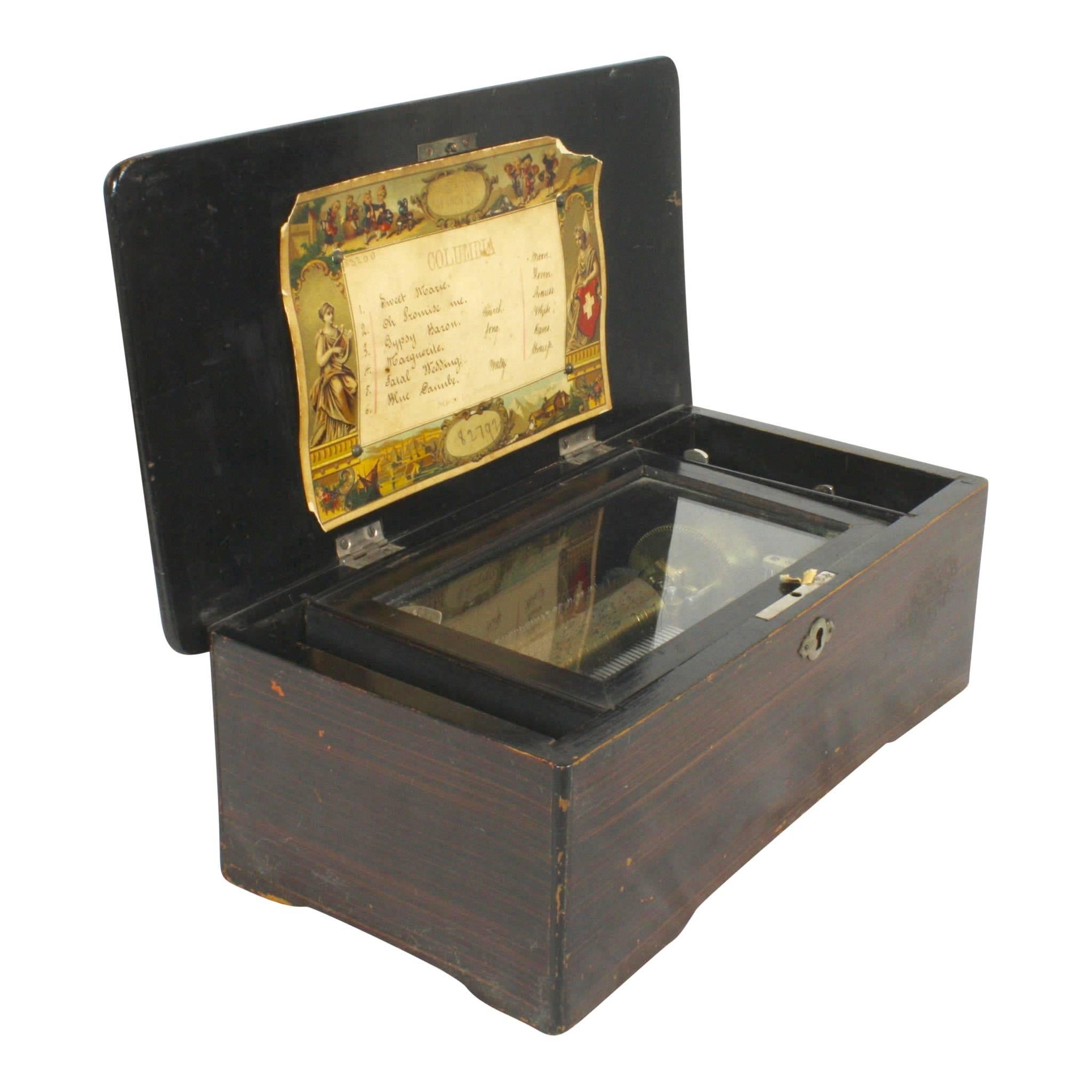 In good working condition, this Swiss music box plays six songs, which are listed under the Columbia label inside the lid. When cranked, music is produced as the pinned, brass cylinder is struck by the comb. The hand crank is removable and can be