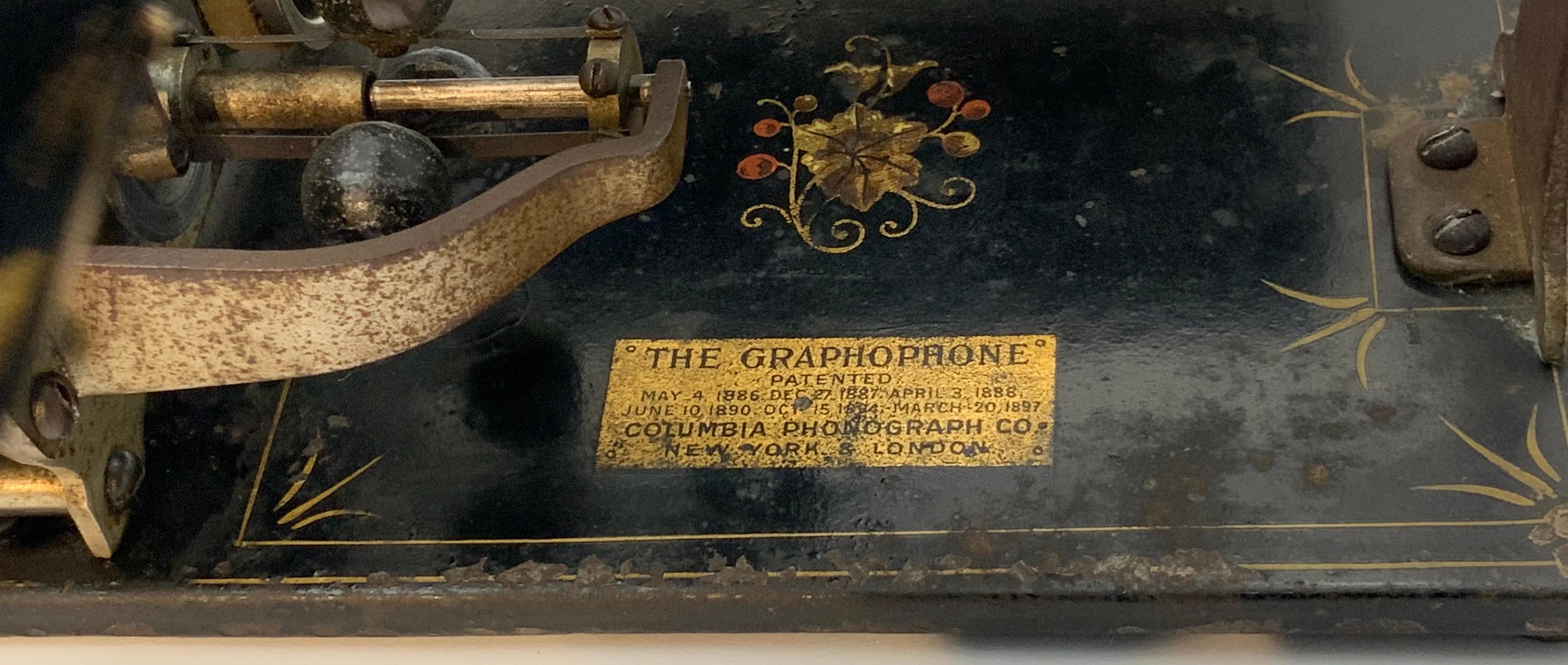 A Graphophone model Q with a black horn adorned with a gold line. It stands over a black iron base decorated with grapevine decal & gold accents. Manufacturer’s name and addresses are impressed in the front of the base. Patents dates are incised in