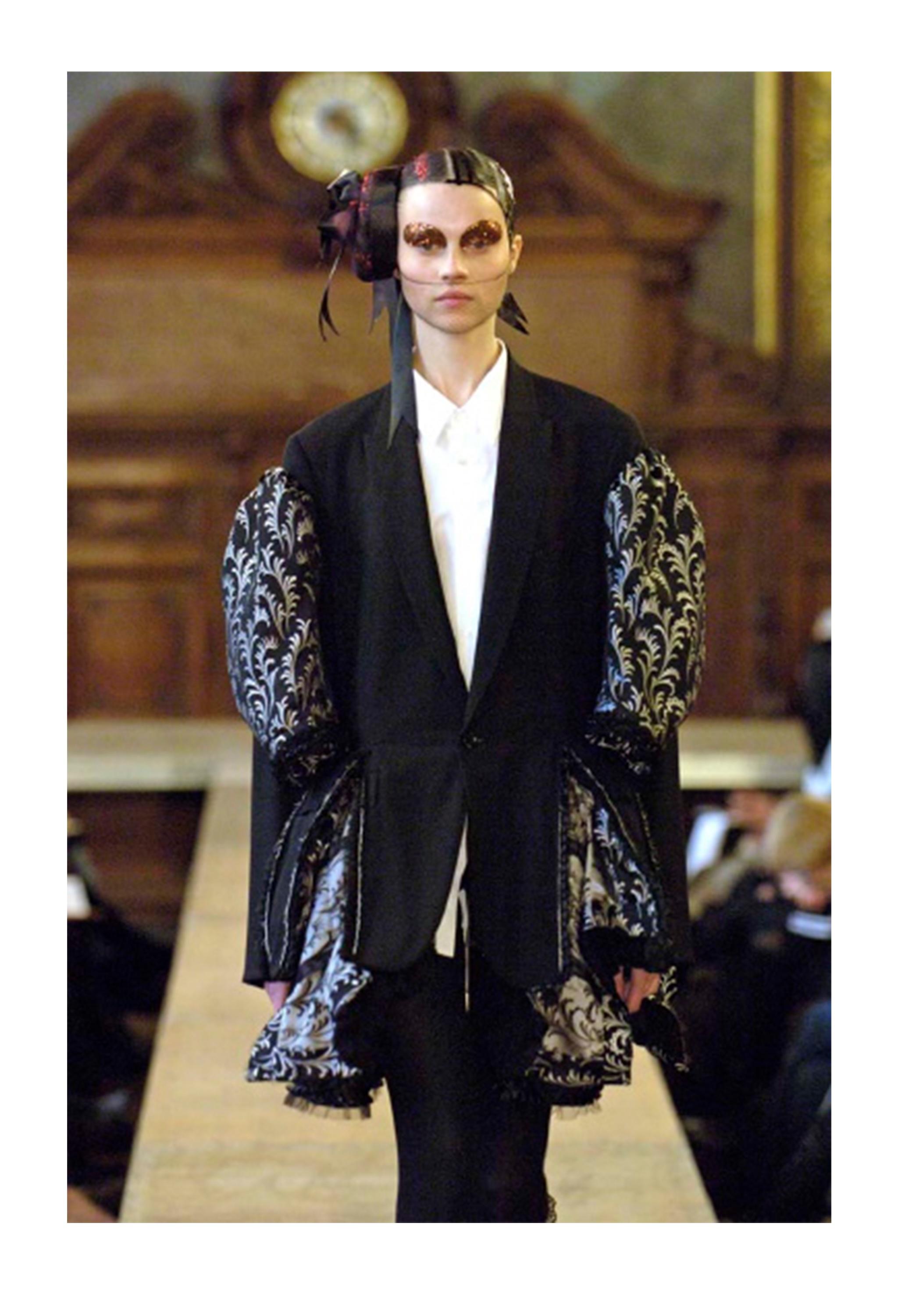 Resurrection Vintage a le plaisir de vous proposer un manteau vintage noir Comme des Garçons avec des broderies argentées, des manches volumineuses à motifs, un ourlet à volants en forme de péplum et une fermeture à bouton unique.

Comme Des