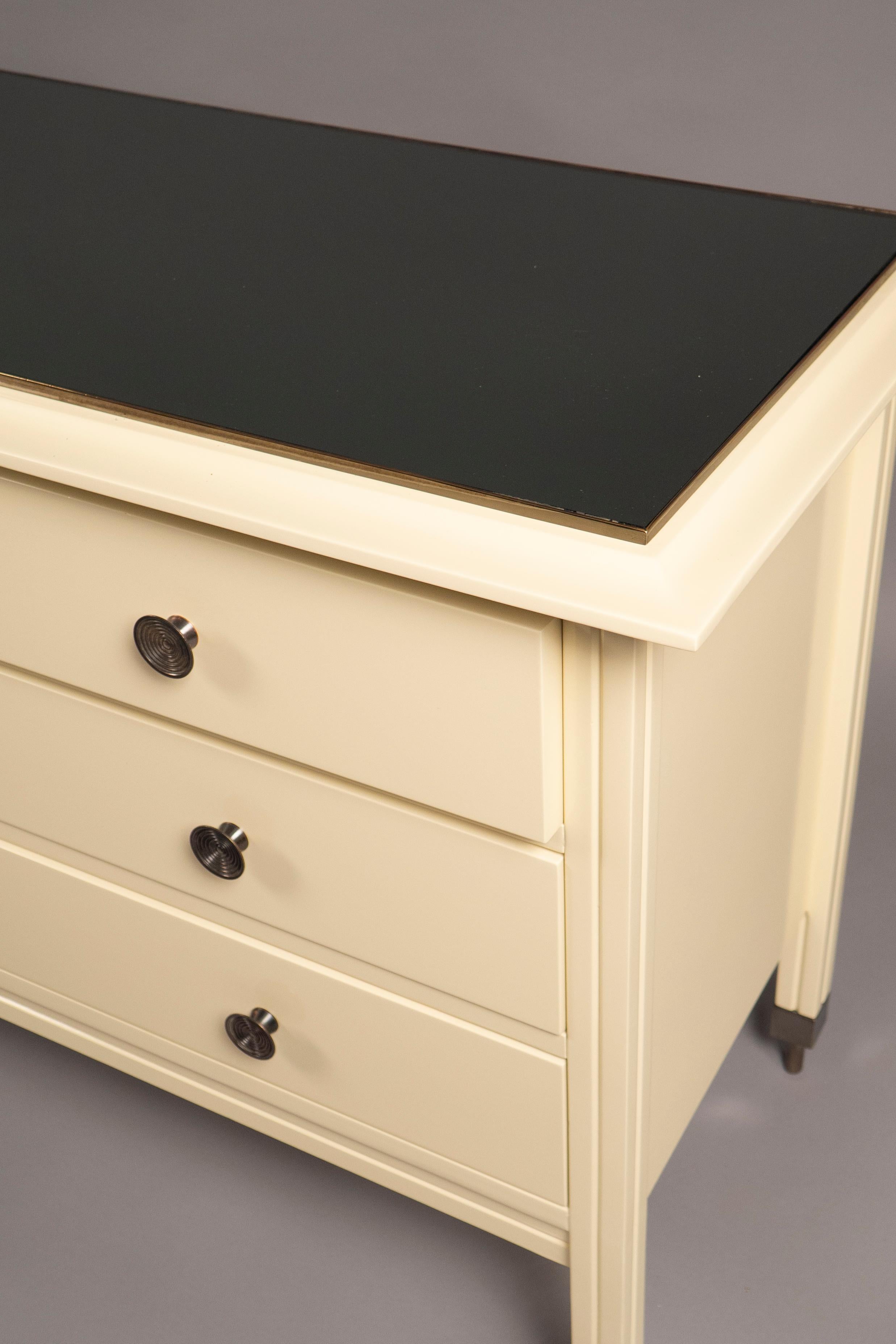 Off-white lacquered wood three drawer chest, fitted with a dark mirrored glass top and patinated pulls, raised on four square patinated legs; produced by Sormani, model D 154.
 