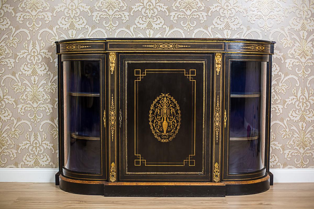 We present you this piece of furniture, circa 1850, painted black, with intarsia’s made of a light type of wood.
This commode is three-door, with a solid central door, and glazed corners in the semi-circular shape.
The frame of the central door