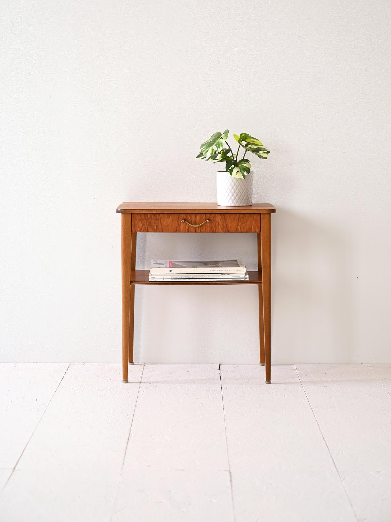 Comodino vintage anni '60 in teak, dotato di un cassetto con impugnatura in metallo e un piano portariviste, è un elegante esempio di design versatile e funzionale.

Questo comodino si distingue per la sua struttura in teak, che conferisce un caldo