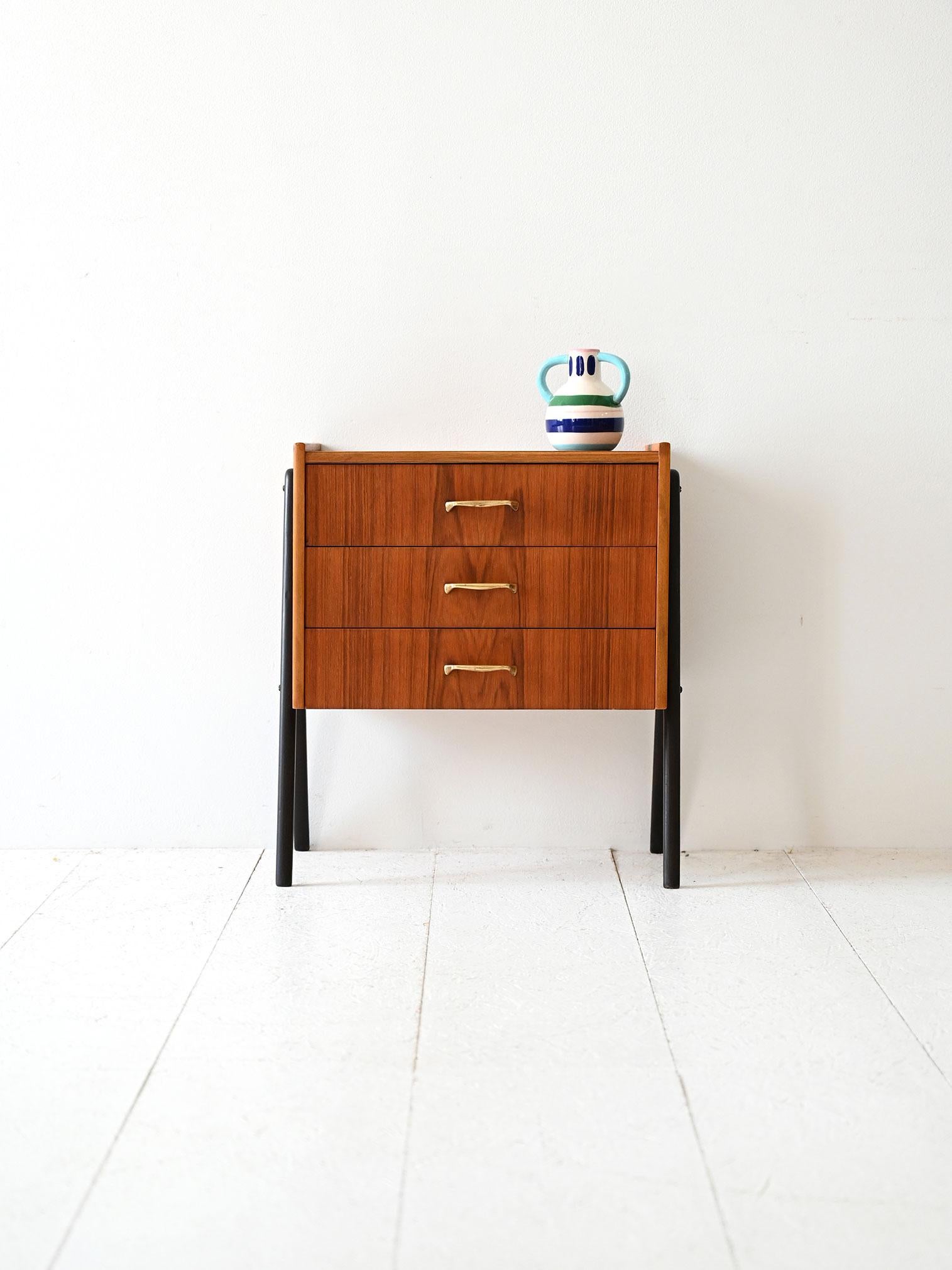 Comodino vintage in teak con tre cassetti, maniglie in ottone e gambe nere inclinate dal profilo grafico.

Comodino scandinavo originale degli anni ’50, realizzato in teak con tre cassetti dal fronte lineare e maniglie in ottone sagomato. La