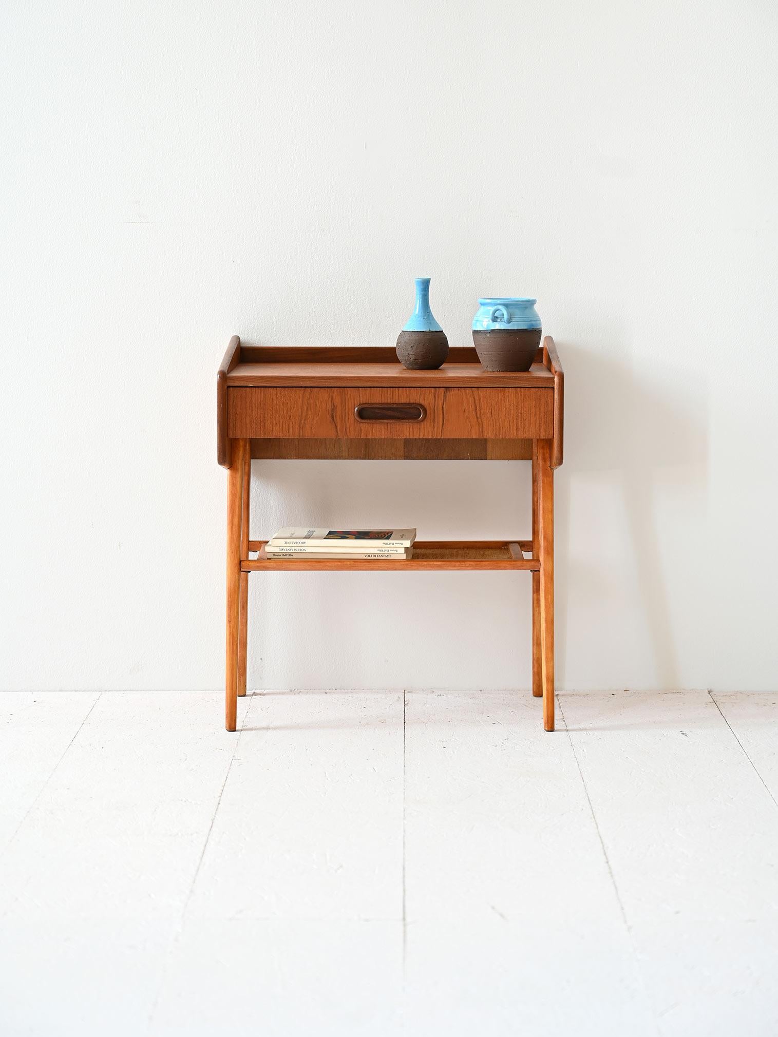 Fabriquée en bois de teck fin, cette table de chevet incarne le style distinctif de l'époque. Le tiroir, doté d'une poignée en bois sculpté, offre un espace de rangement pour les petits objets essentiels.

Les pieds élancés, légèrement inclinés vers