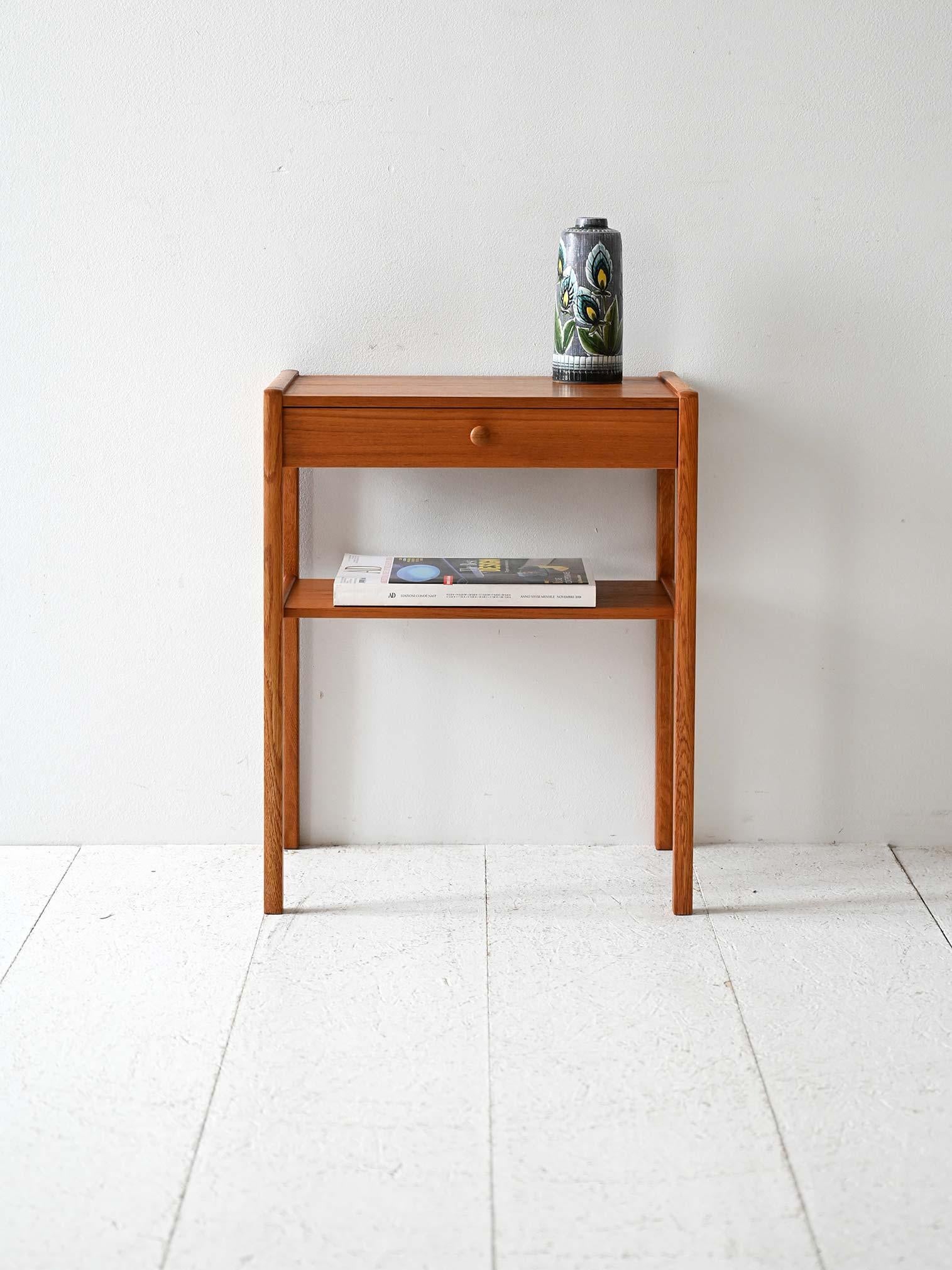 Comodino in teak con cassetto centrale e ripiano basso. Linea semplice e struttura snella.

Comodino scandinavo vintage in teak con struttura essenziale. Il cassetto superiore ha una maniglia tonda in legno integrata, mentre la parte inferiore