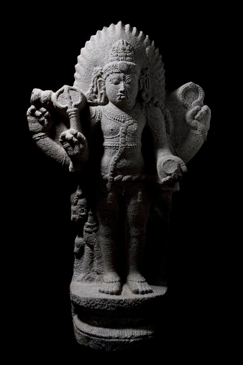 Complete granite figure of Shiva showing his attributes in a standing hieratic pose, fully dressed and jeweled in royal regalia with tiara on a high pedestal with a flaming halo. Late Vijayanagar Dynasty.