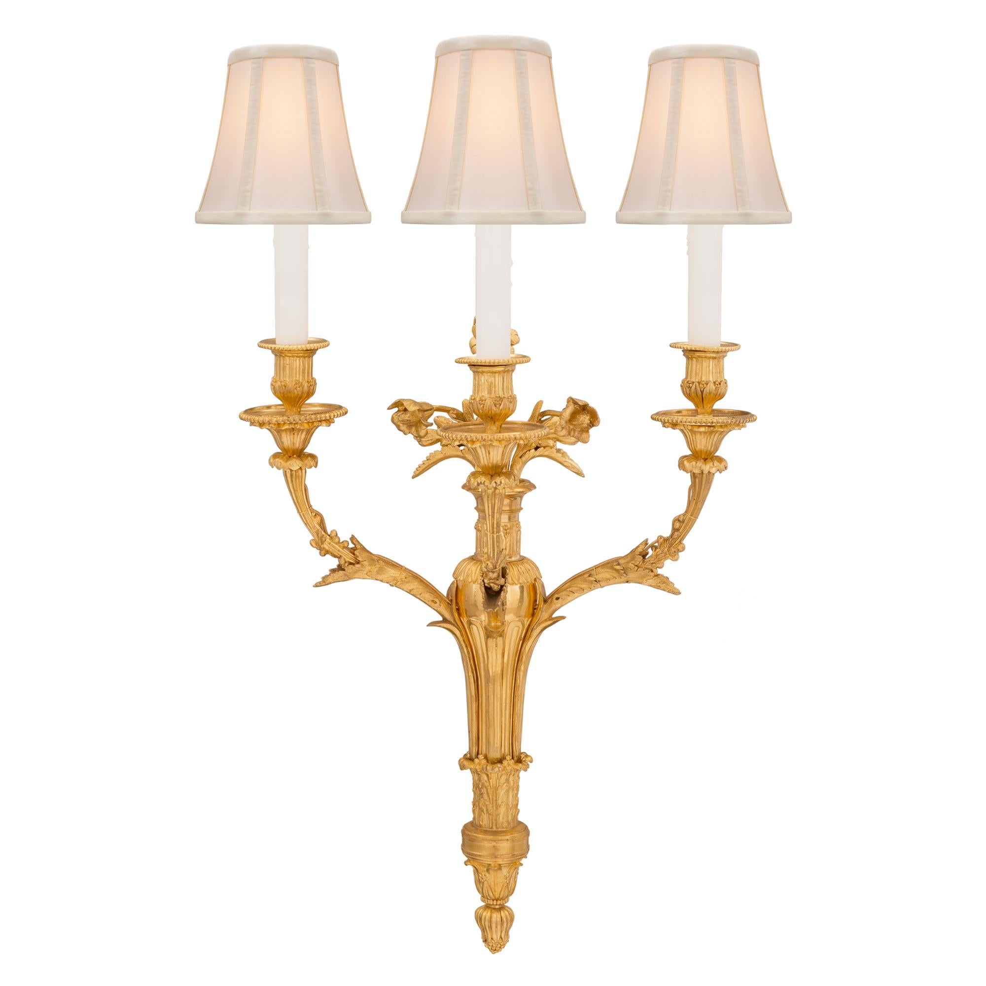 A stunning and extremely high quality complete set of six French 19th century Louis XVI St. ormolu sconces. Each three arm sconce is centered by a lovely bottom floral acorn finial below the elegant fluted lightly tapered central support adorned