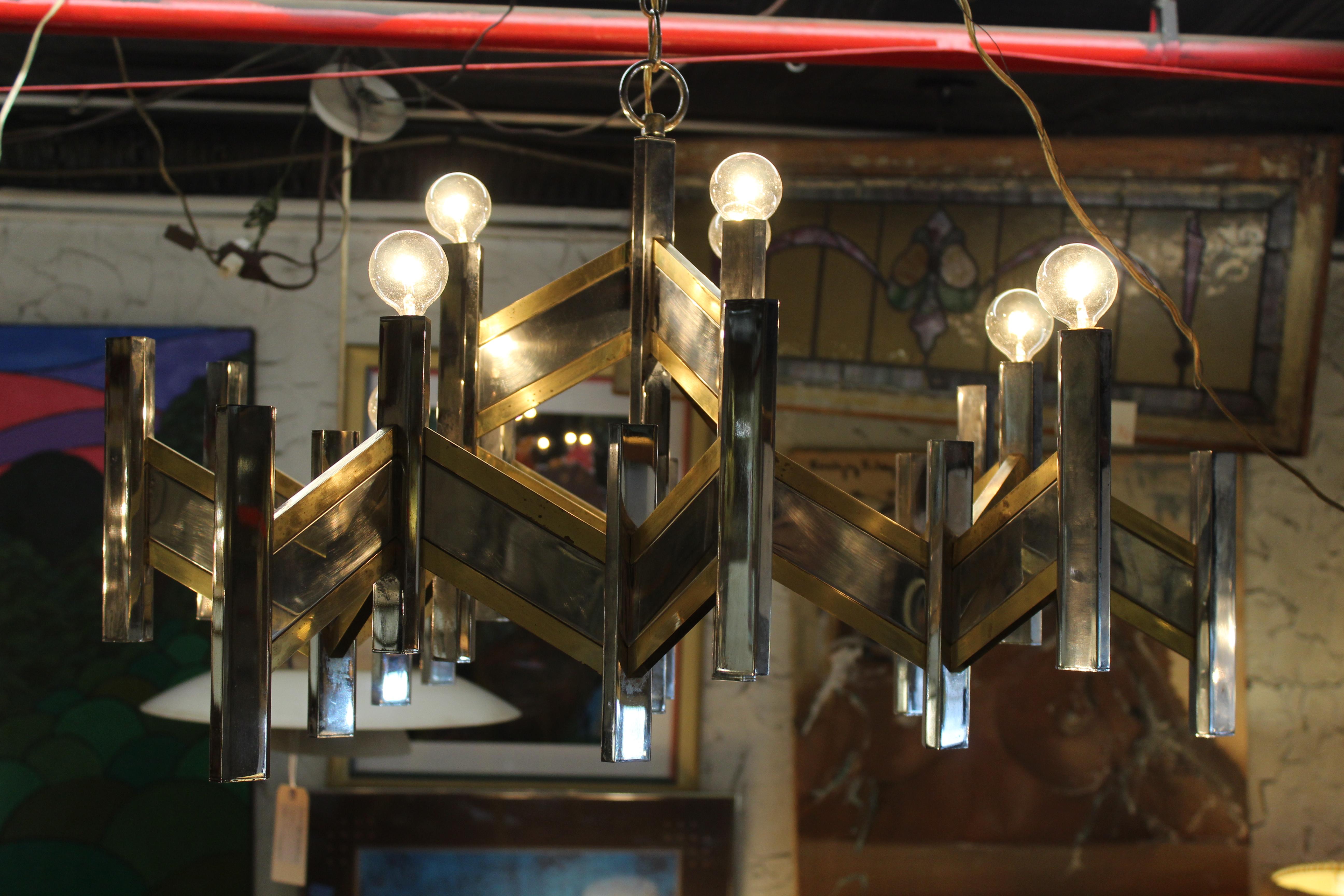 Lampadario geometrico e distintivo realizzato in metallo con eleganti finiture in ottone. L'audace design angolare fonde una struttura modernista con caldi dettagli metallici, creando un punto focale di grande impatto per sale da pranzo, ingressi o