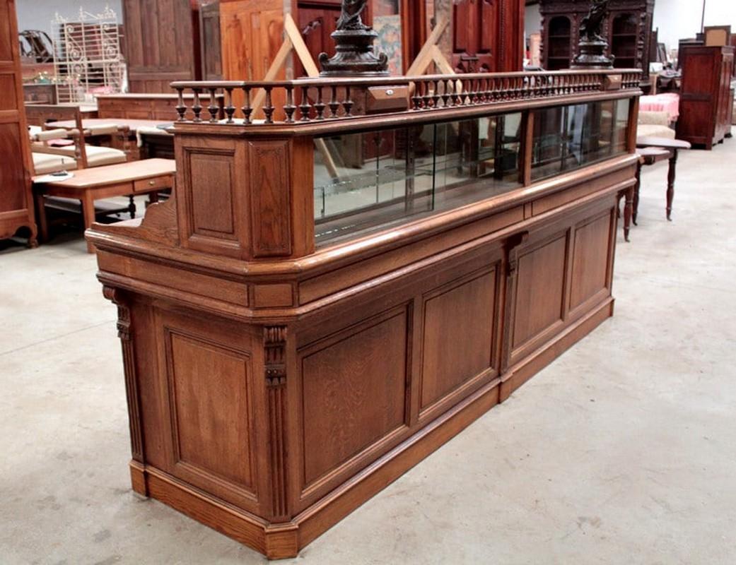 Very large oak pharmacy counter opening on the front with four sliding glass doors and a small glass shelf on a mirrored background in the upper section, topped with a baluster gallery. The lower section is composed of four oak panels surrounded by