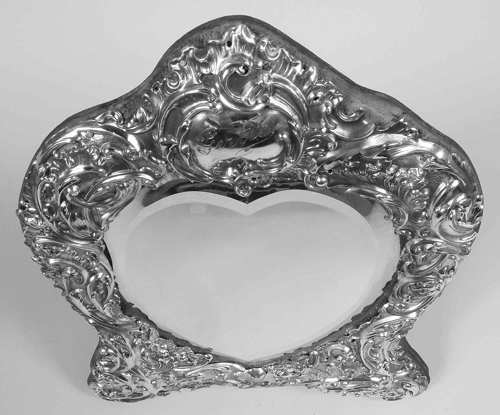 Edwardian Rococo sterling silver mirror. Made by William Comyns in London in 1907. Heart-shaped beveled glass in shaped surround with pointed top and bracket supports. Embossed leafing scrolls and flowers in pell-mell, dynamic arrangement. At top