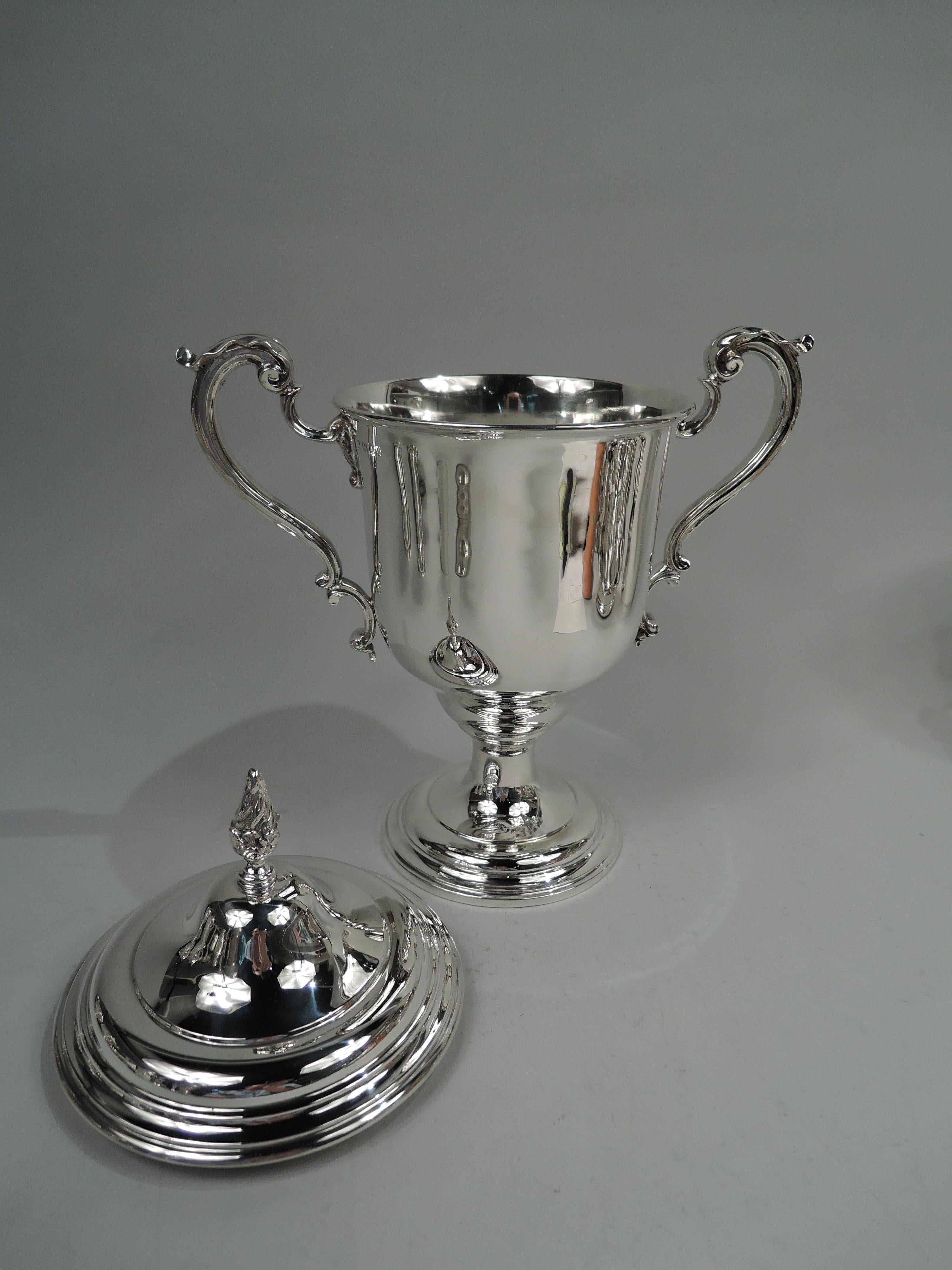 George V sterling silver trophy cup. Made by Charles & Richard Comyns in London in 1922. Urn with ovoid bottom on raised and stepped foot; leaf-capped high-looping double-scroll side handles. Cover stepped and domed with cast flame finial.