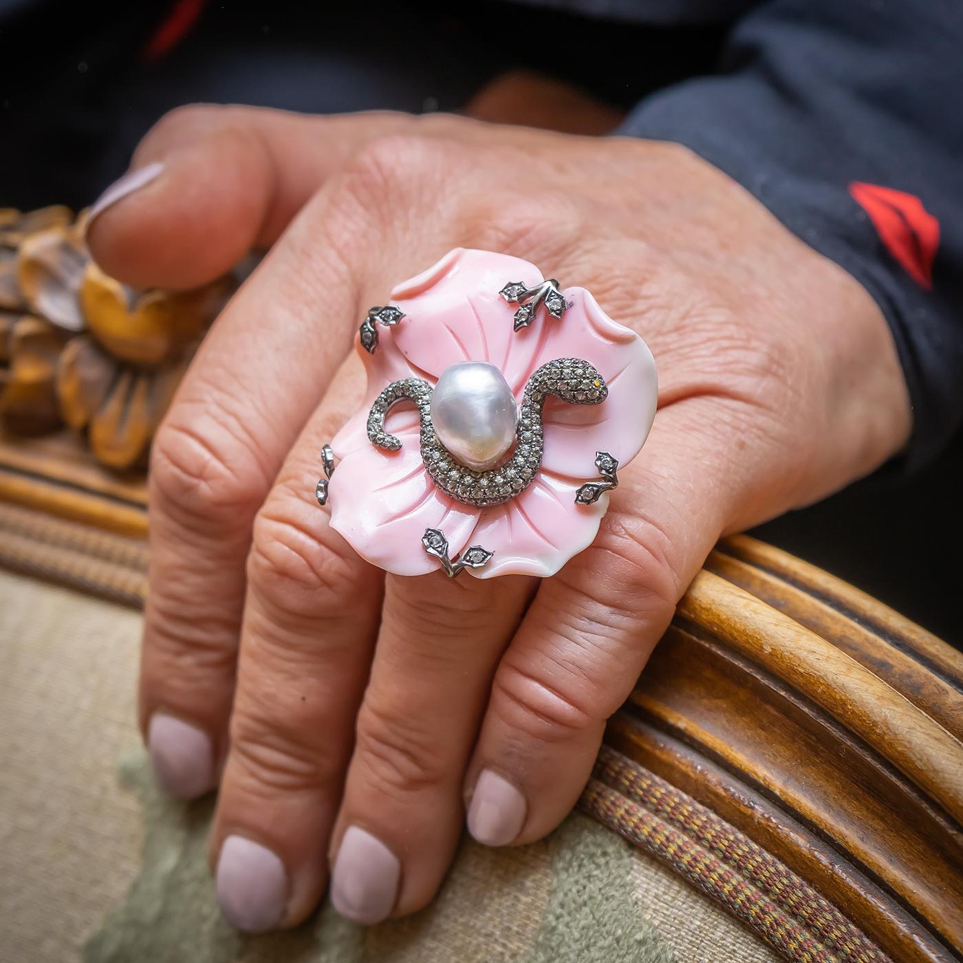 A carved, queen conch shell, flower and snake ring, with a carved, pink conch, shell flower, with a grey pearl, measuring 13 x 10mm, with a snake, curling around the central pearl, pavé set with grey sapphires, and small diamond set leaves, mounted