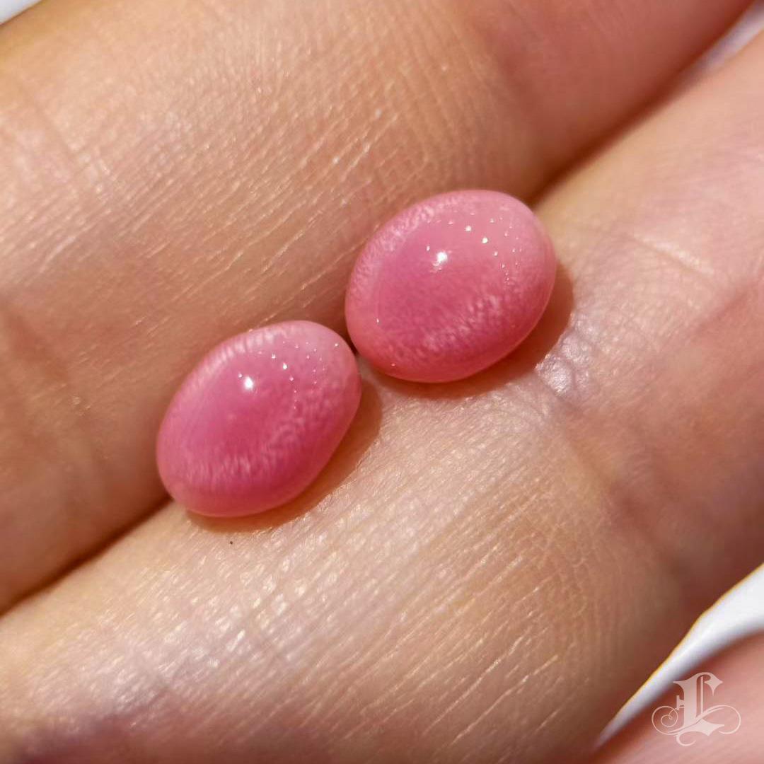 A rare matched pair of natural conch pearls, glowing with saturated flamingo pink and pronounced flame patterning. Measuring 7.8 x 5.6 mm and weighing 1.68 + 1.50 carats, these oval cabochons are ideal for crafting a stunning pair of earrings. Their