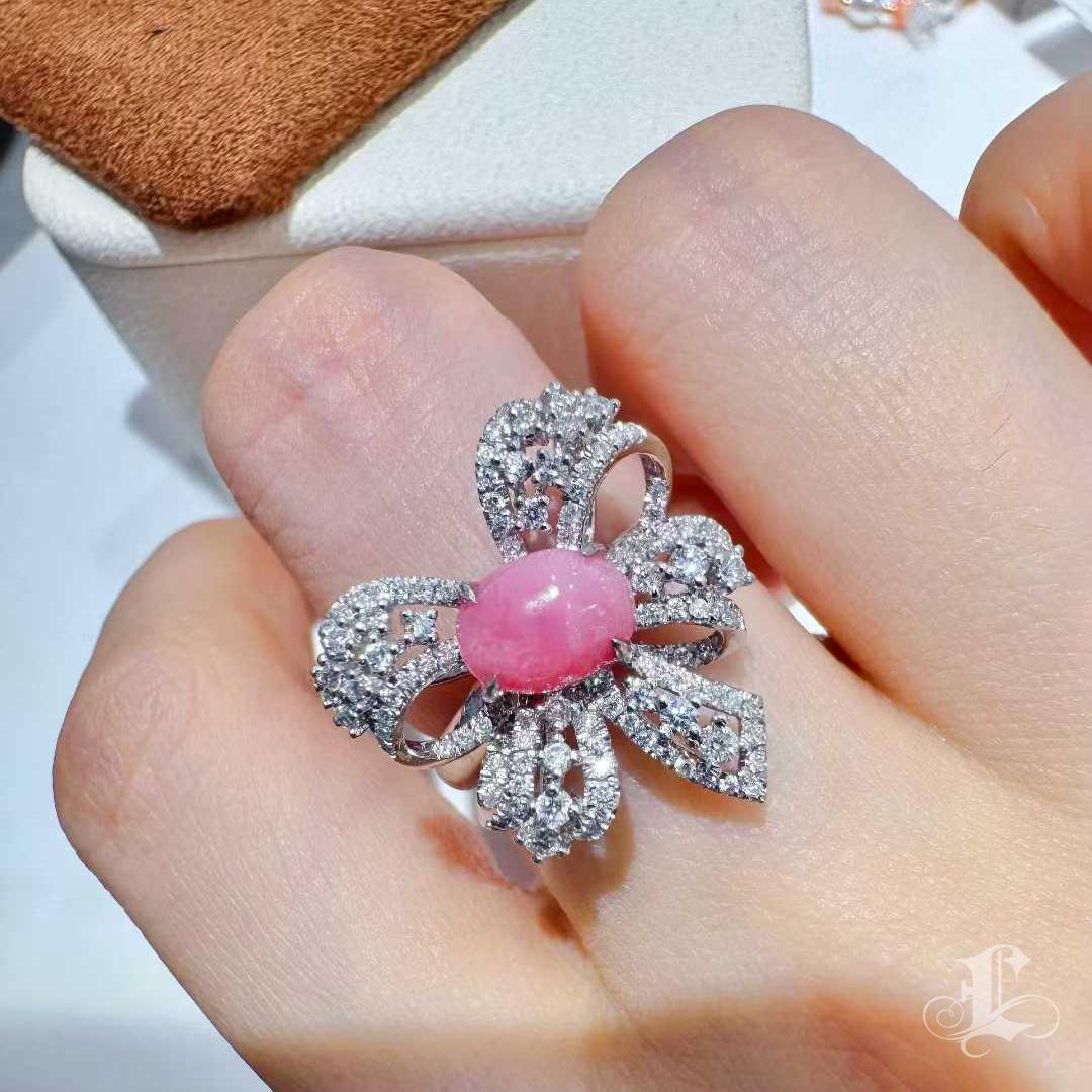 A rare and exquisite ring featuring a 1.46ct natural conch pearl in a delightful flamingo-pink hue, displaying a pronounced flame pattern unique to conch pearls. The gem’s oval and well-proportioned shape is embraced by a graceful diamond bow