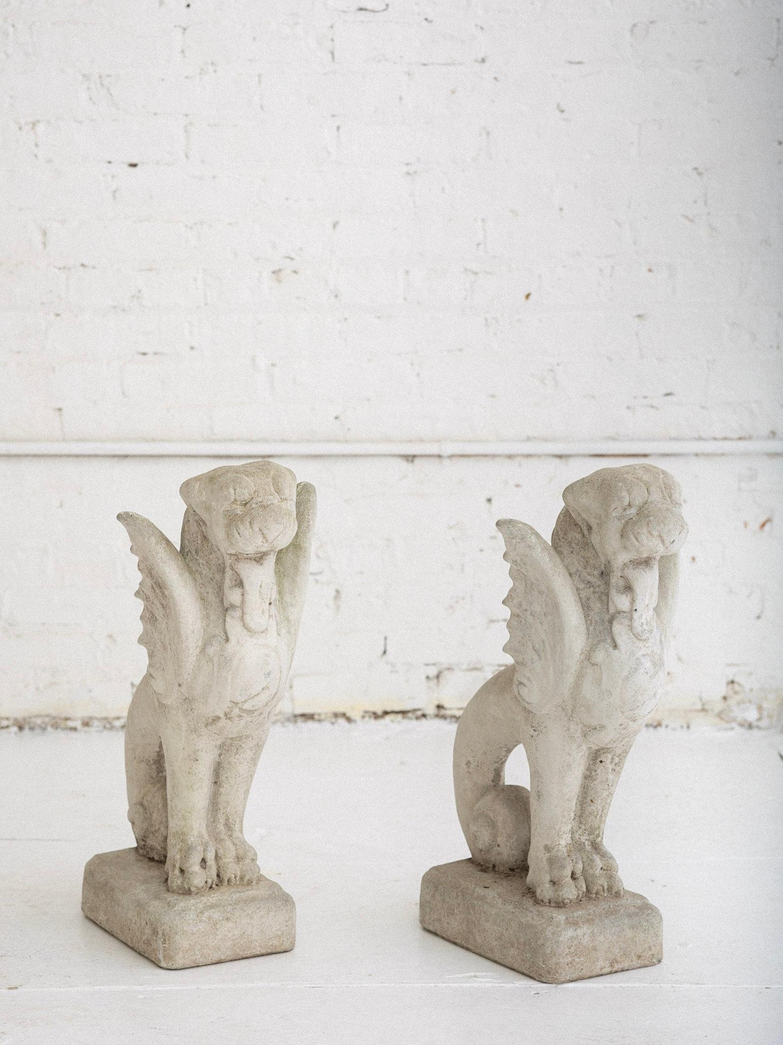 A pair of concrete cast stone garden statues. Mythical creatures with a dog or lion face, wings, fish tail and shield. Reminiscent of a gargoyle. An outdoor accent for a garden or landscaping. Weathered patina.